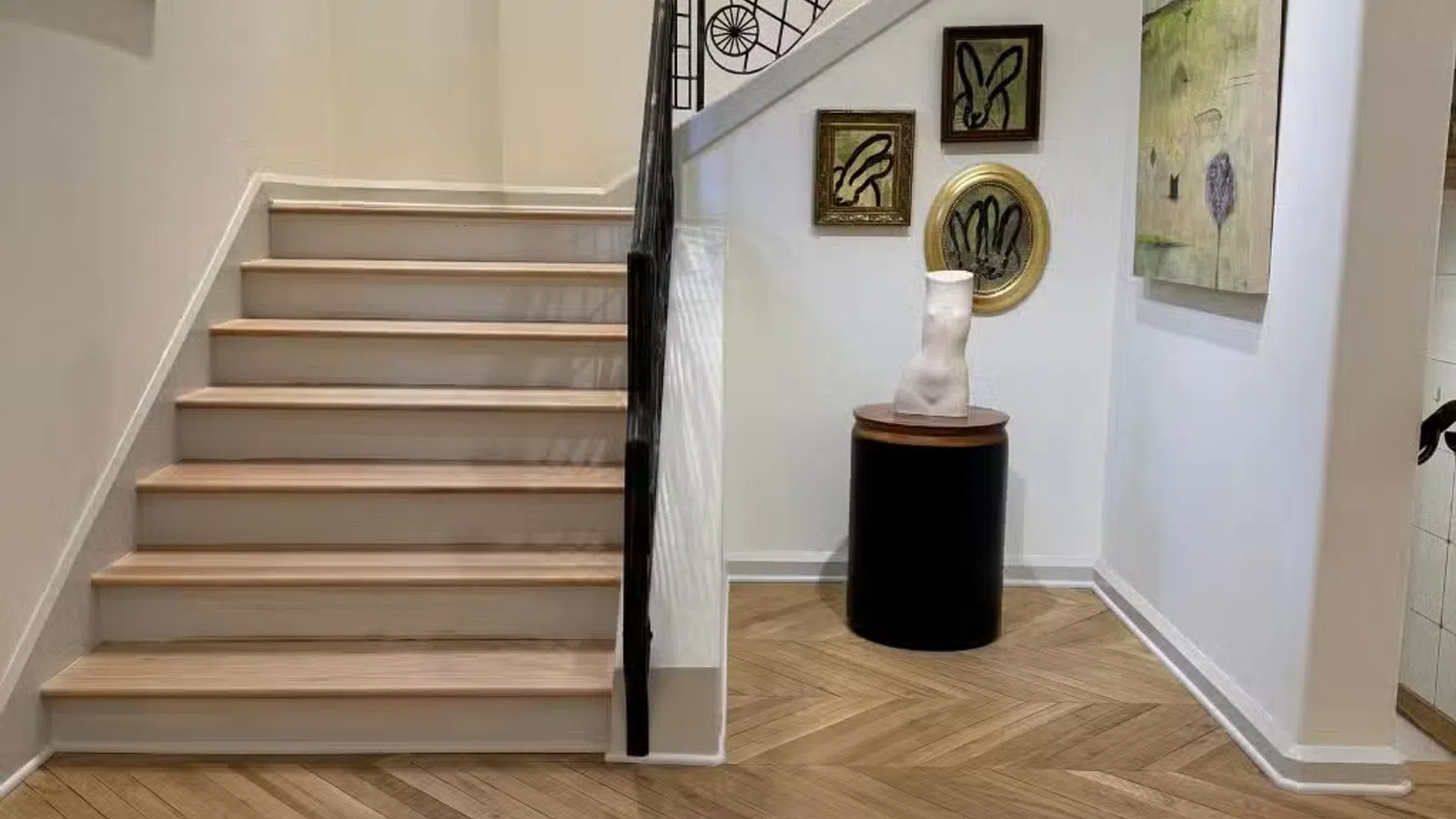 Modern interior with wooden herringbone floor, staircase, decorative sculpture, and framed artwork on white walls.