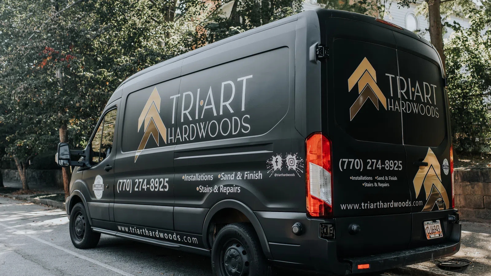 Black service van featuring Tri-Art Hardwoods logo, phone number, website, and service details parked on street.