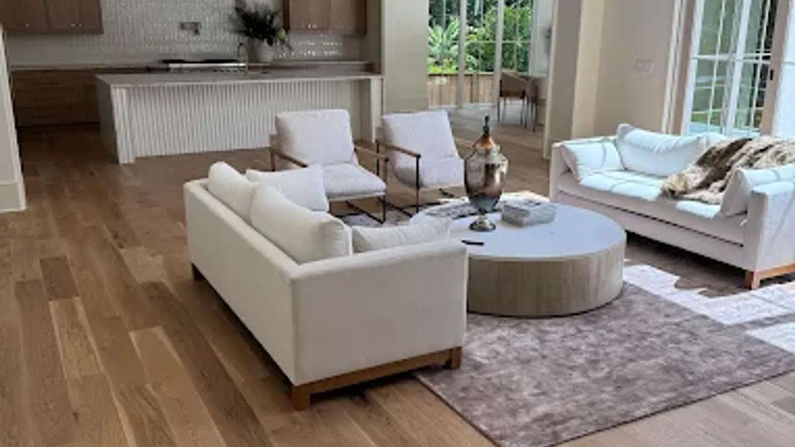 Spacious modern living room with white sofas, armchairs, wooden flooring, and an open kitchen in the background.