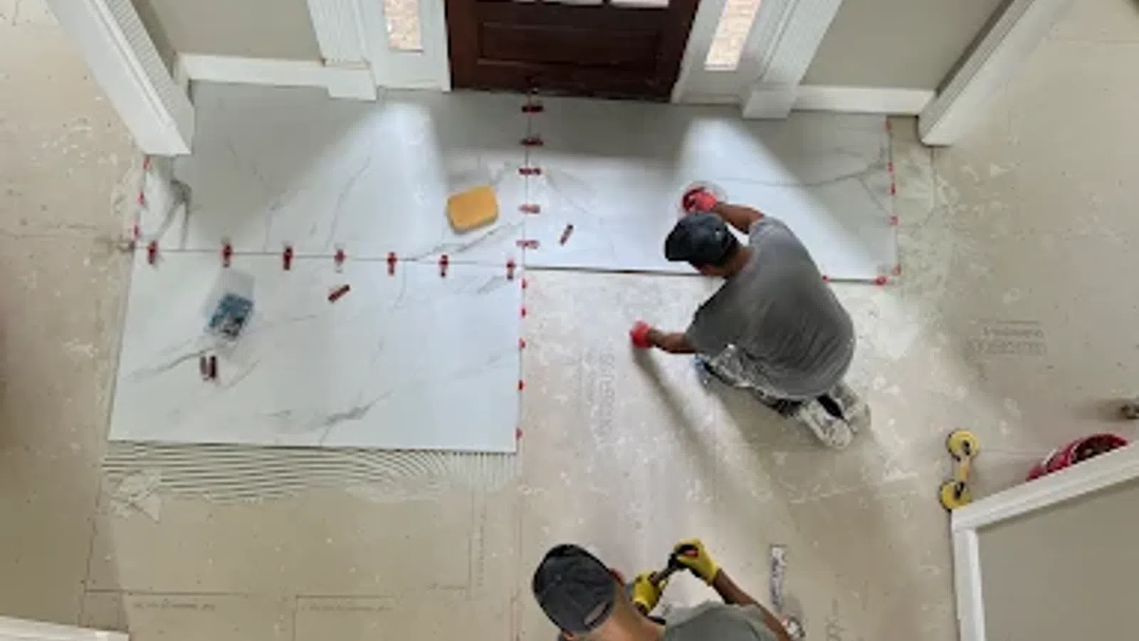 Two workers installing large white marble floor tiles near a wooden door inside a house under construction.