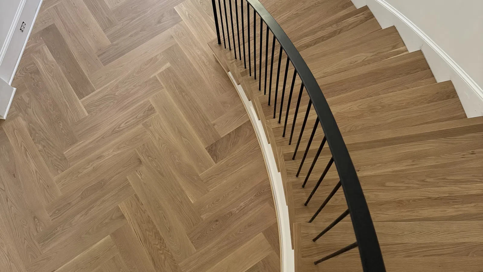 Curved wooden staircase with black railing and chevron-patterned wood flooring in entryway with dark door.