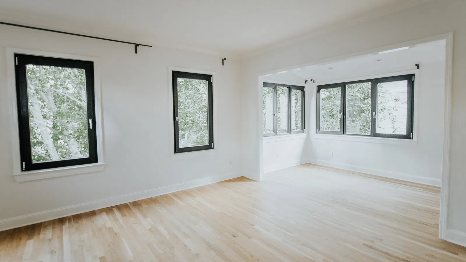 Bright empty room with light hardwood floors and large black-framed windows overlooking green trees.