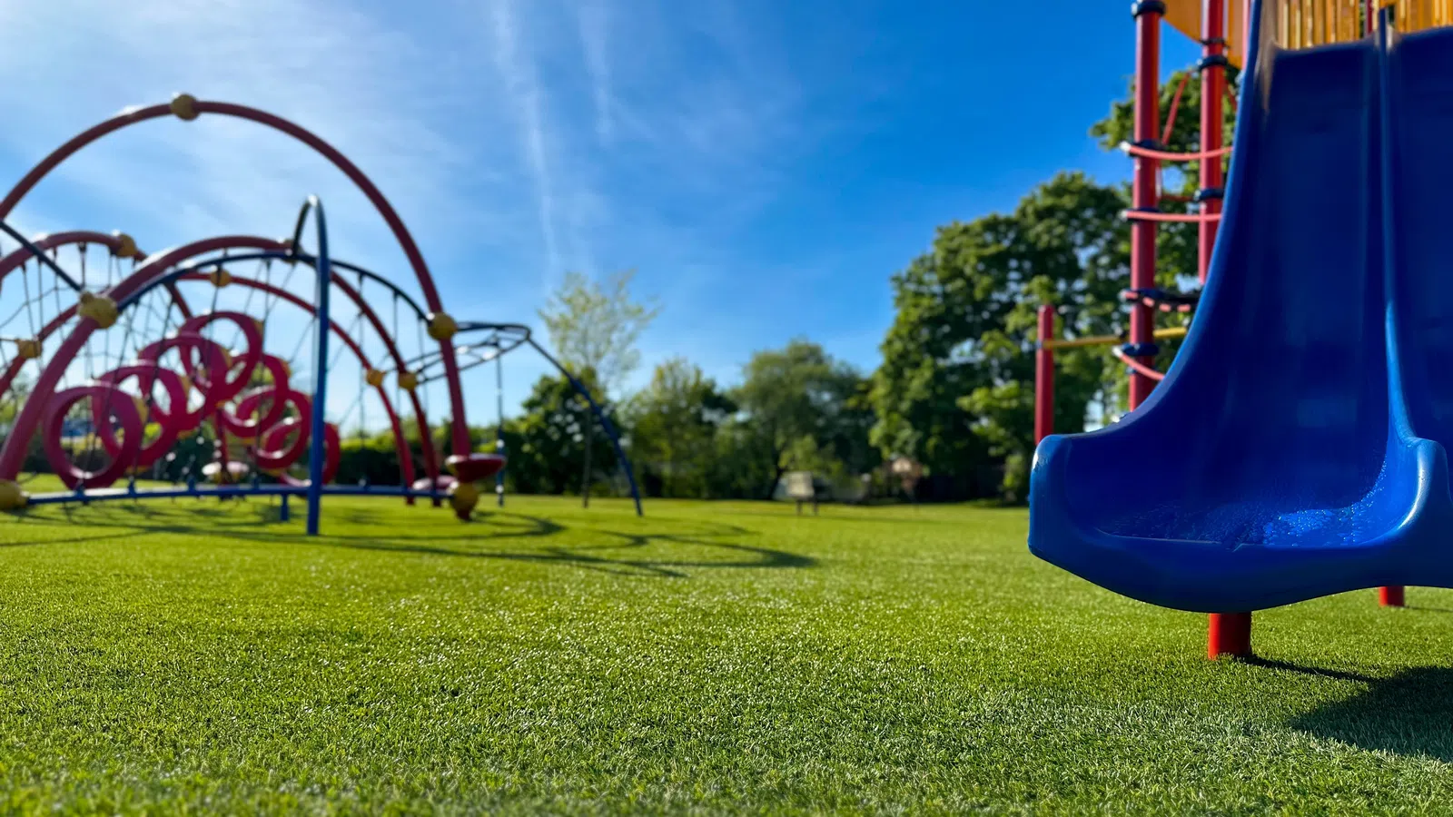 Bright blue sky over a green playground with a blue slide and red climbing structures in a park.