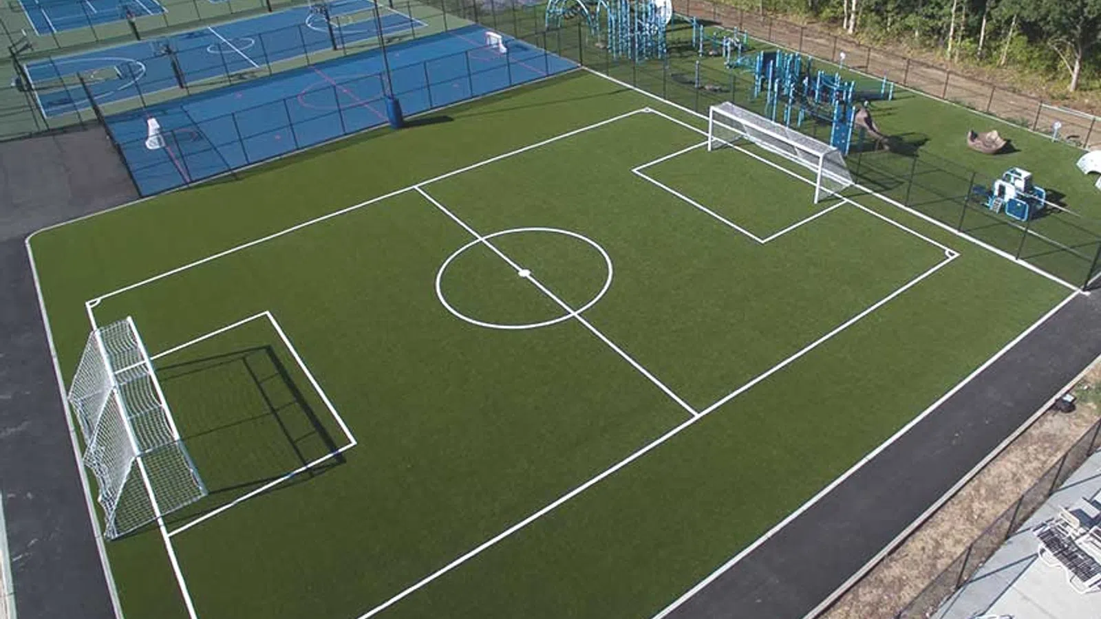 Aerial view of a soccer field with goals, surrounded by tennis courts and playground equipment.