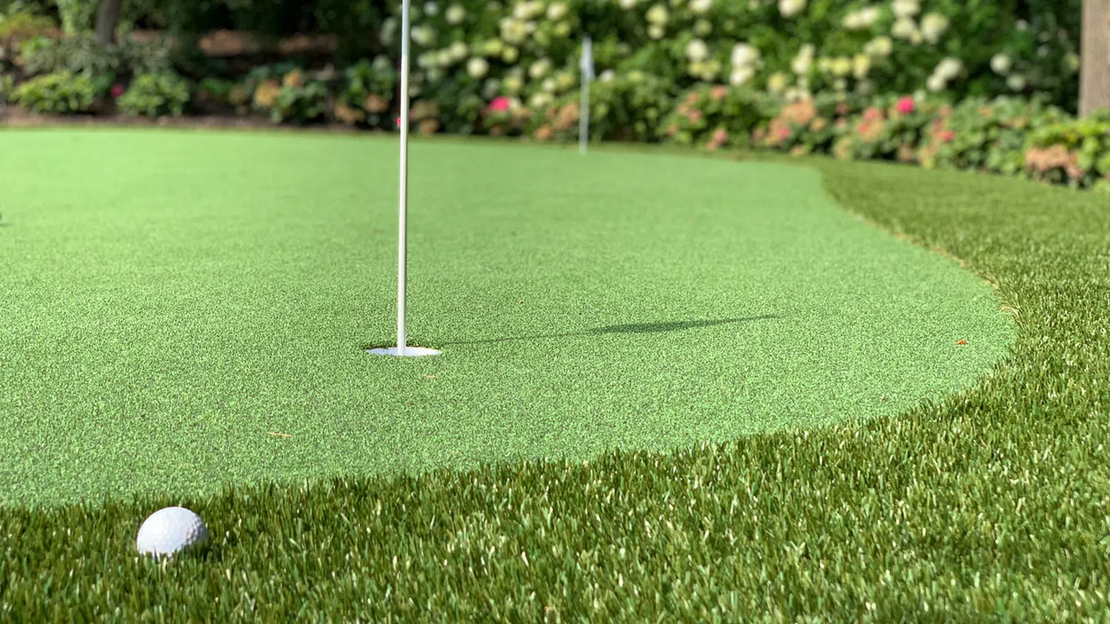 Golf ball near hole on a putting green with flagstick surrounded by garden flowers and grass.