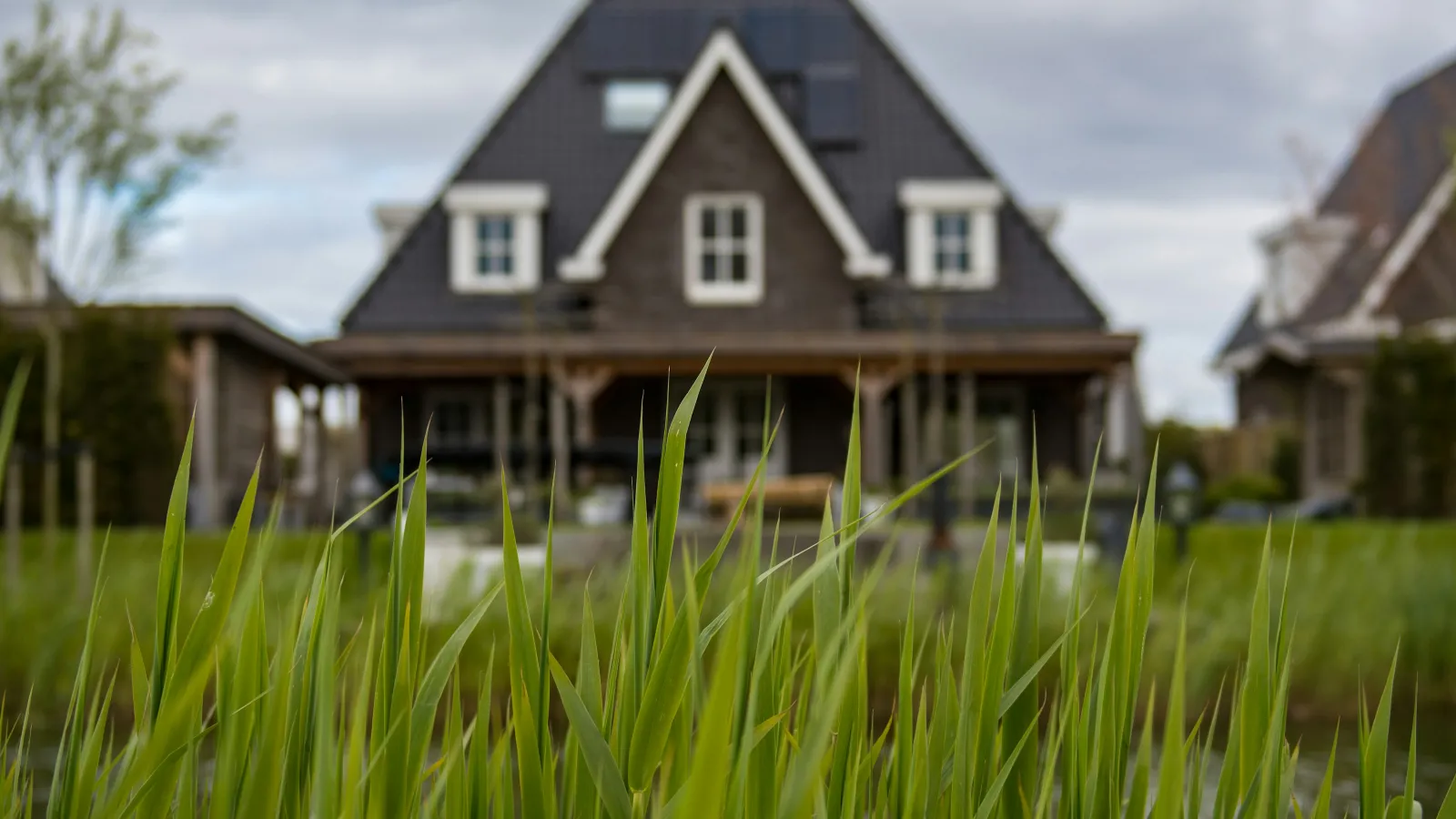A close-up of lush green grass in the foreground with a modern a-frame house in the background under a cloudy sky.