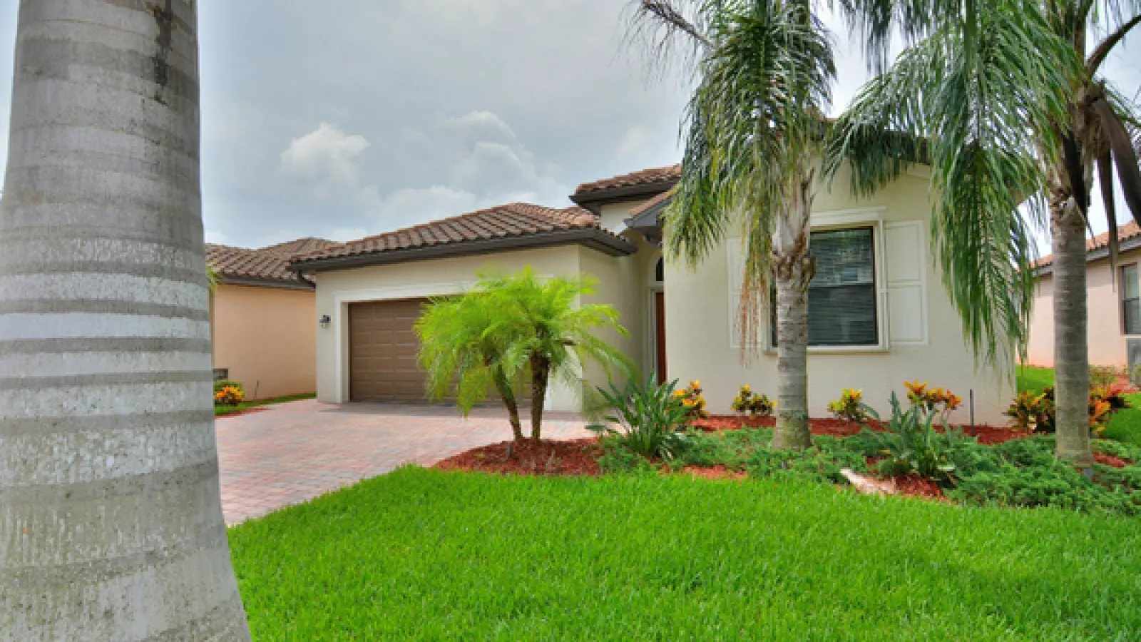 Single-story house with tiled roof, palm trees, green lawn, and brick driveway under cloudy sky