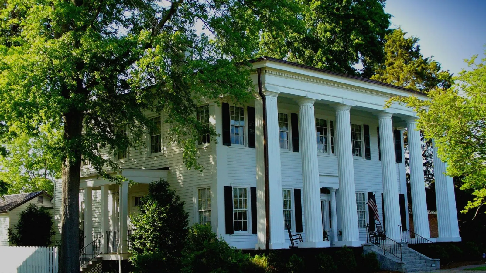 White house with columns surrounded by beautiful large trees and a green, weed free lawn