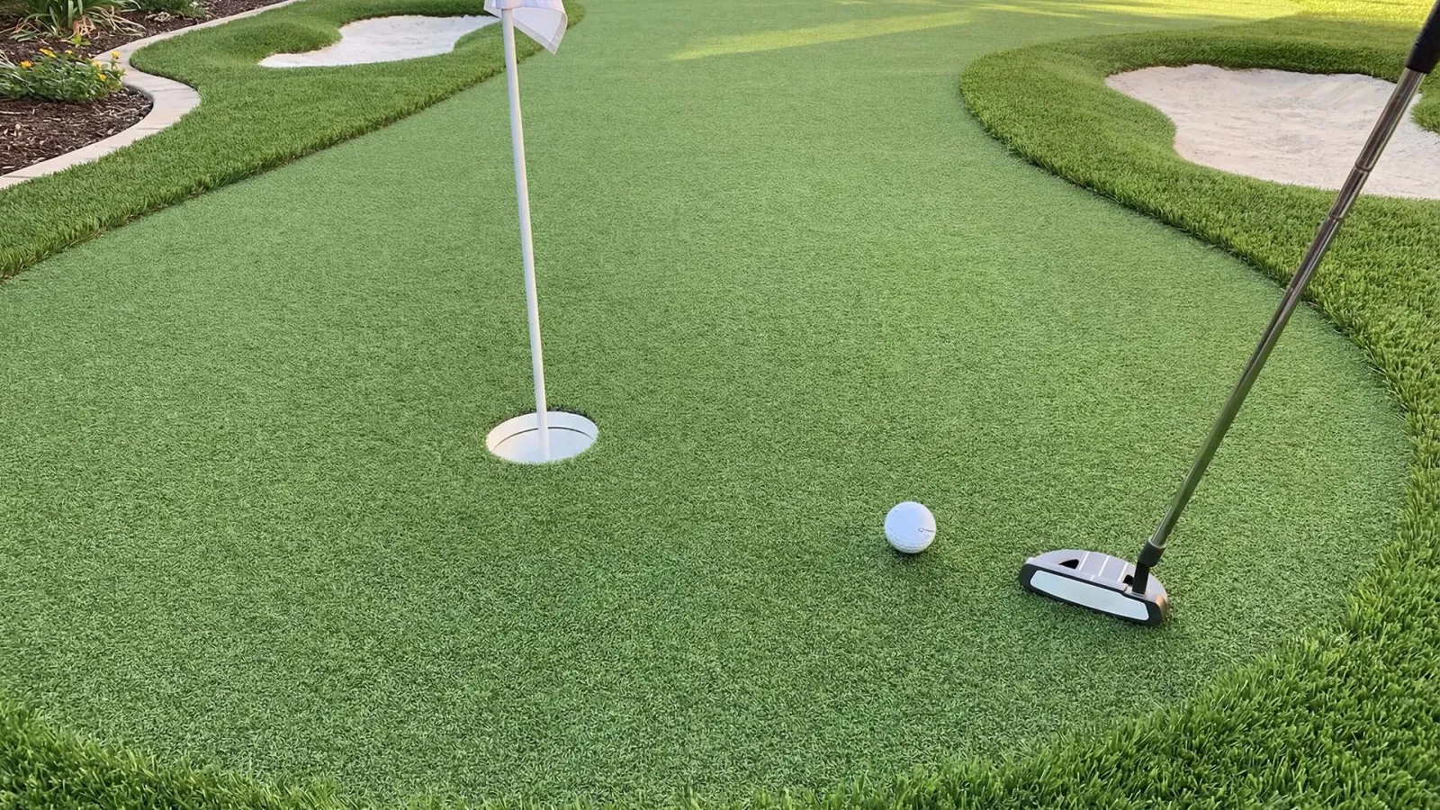 Backyard putting green with two flags, sand bunkers, golf ball, and putter on artificial grass surface.