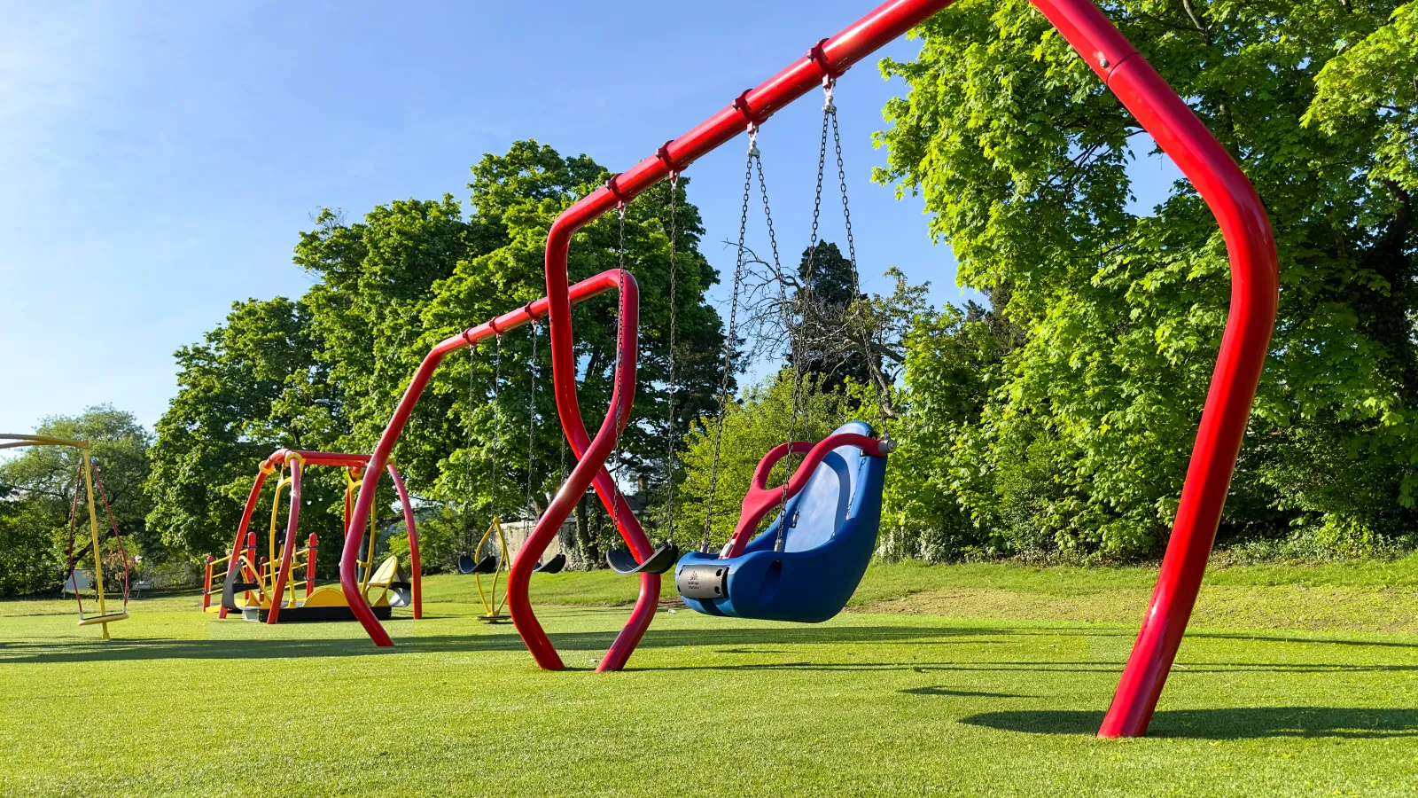 Empty colorful swings with bright red frames on green grass under a clear blue sky at a playground