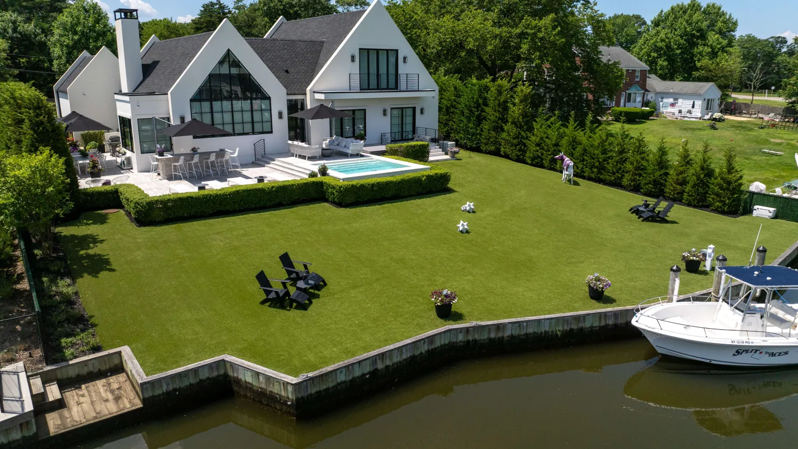 Modern white house with large windows, pool, dock, boat, and manicured lawn by the water on a sunny day