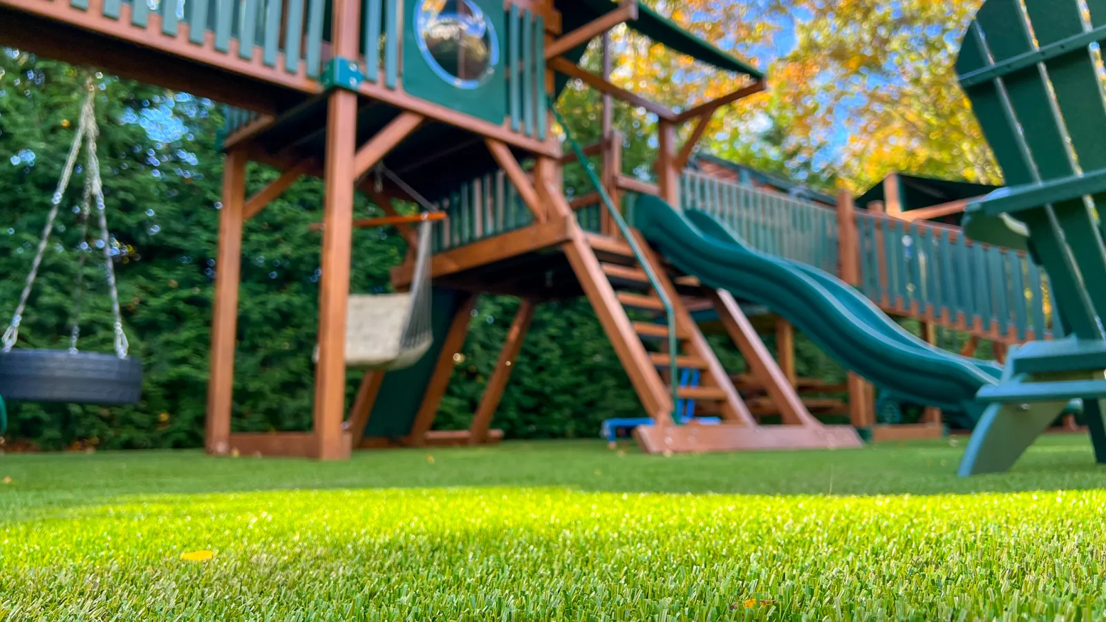 Low-angle view of a backyard playground with wooden playset, green slide, tire swing, and Adirondack chair on green grass.