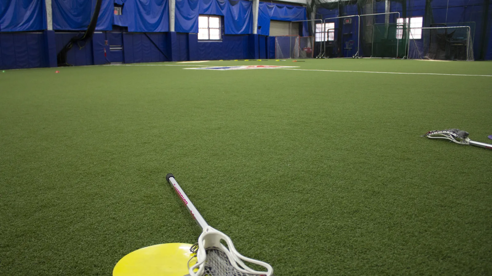 Indoor lacrosse field with artificial turf and lacrosse sticks lying on the ground in a sports facility.