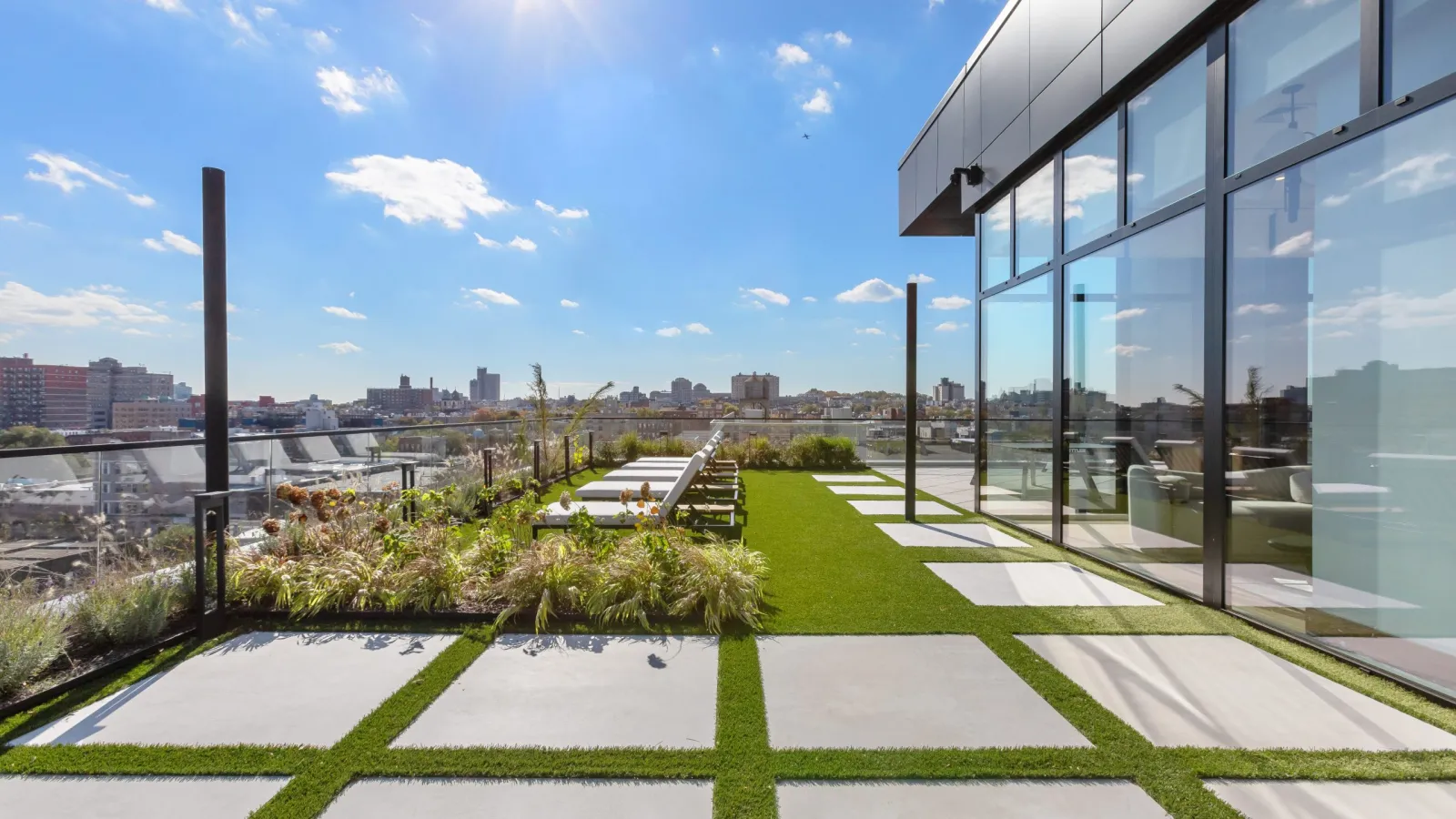 Modern rooftop terrace with concrete pavers, green grass, plants, and city skyline under bright sunlight.