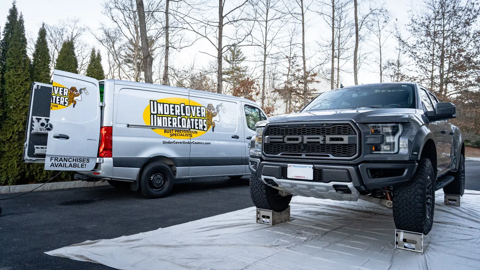 Gray Ford truck elevated on blocks next to UnderCover UnderCoaters van with rust prevention branding.