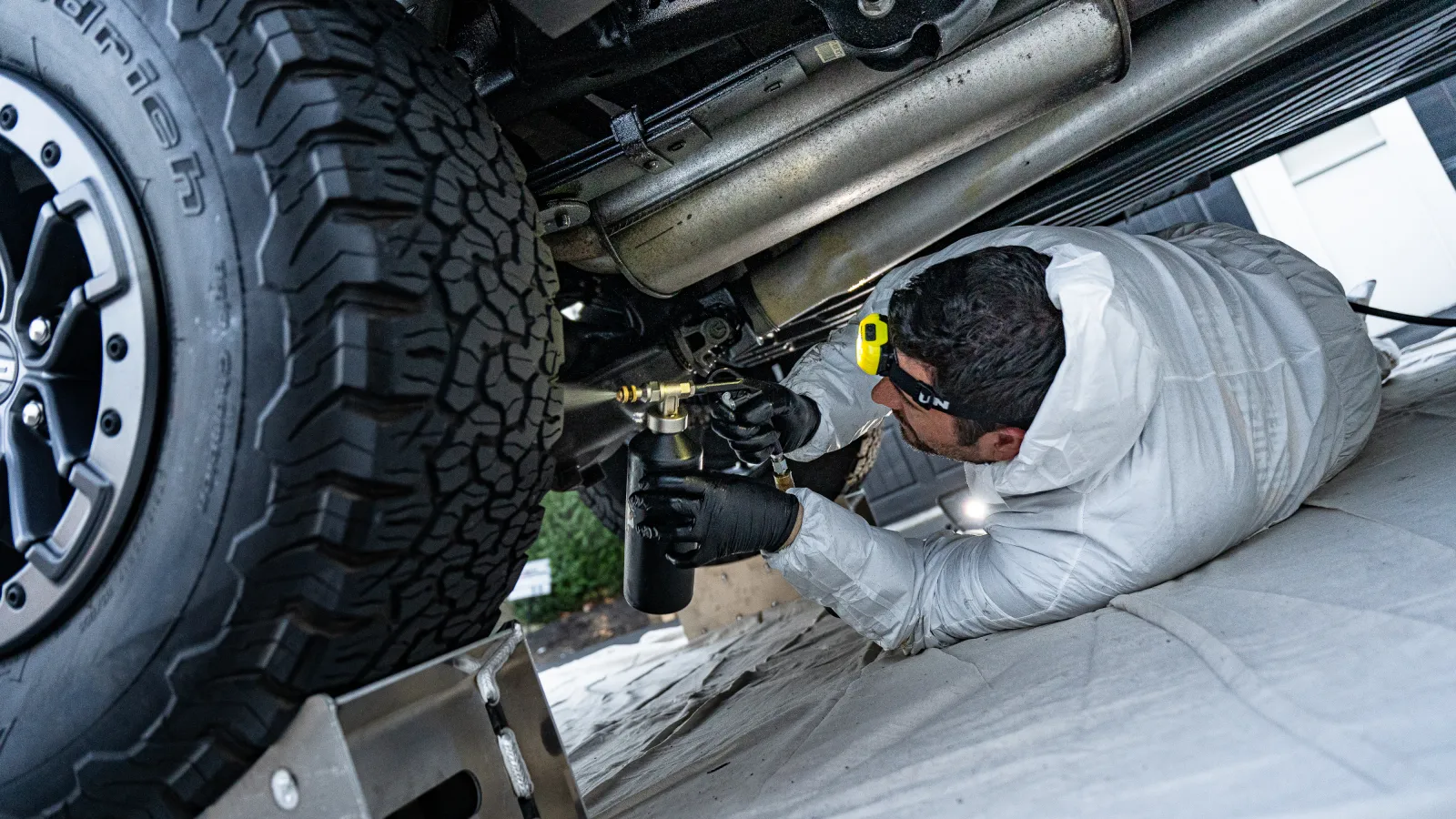 Mechanic in white suit inspecting and repairing vehicle underside near a large tire using specialized tools.