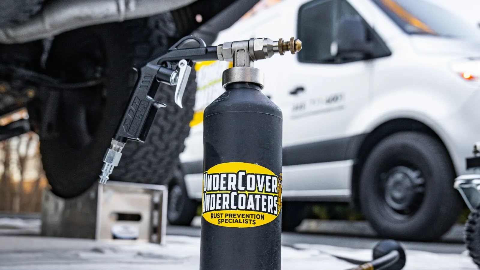 Underbody rust prevention spray canister labeled UnderCover UnderCoaters near lifted vehicle and white van.