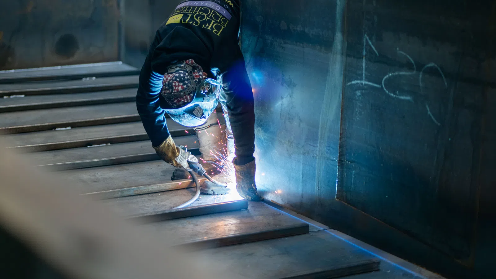 Worker in protective gear welding metal plates inside an industrial workshop with sparks flying.
