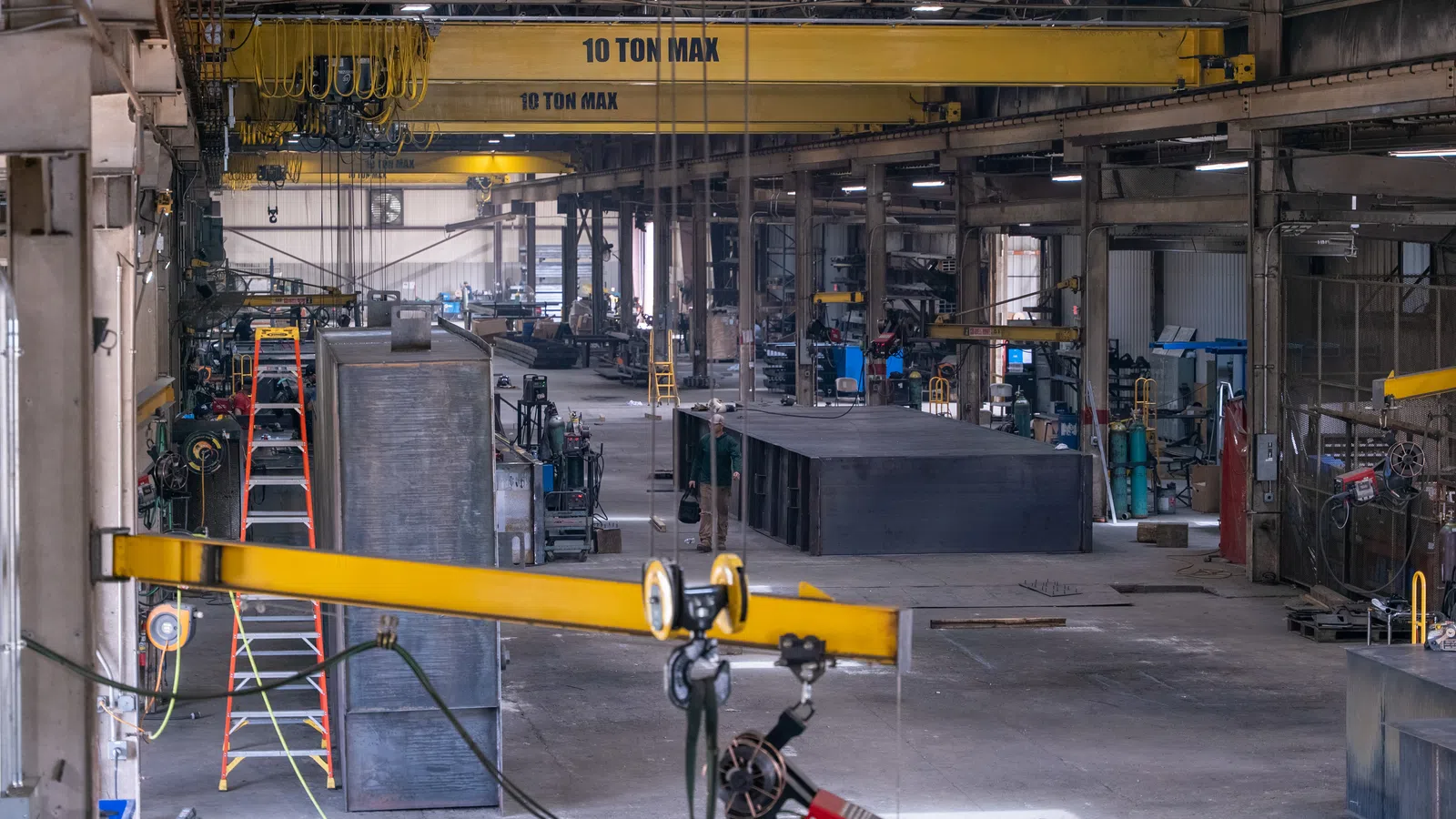 Industrial warehouse interior with overhead cranes, metal structures, ladders, and manufacturing equipment.