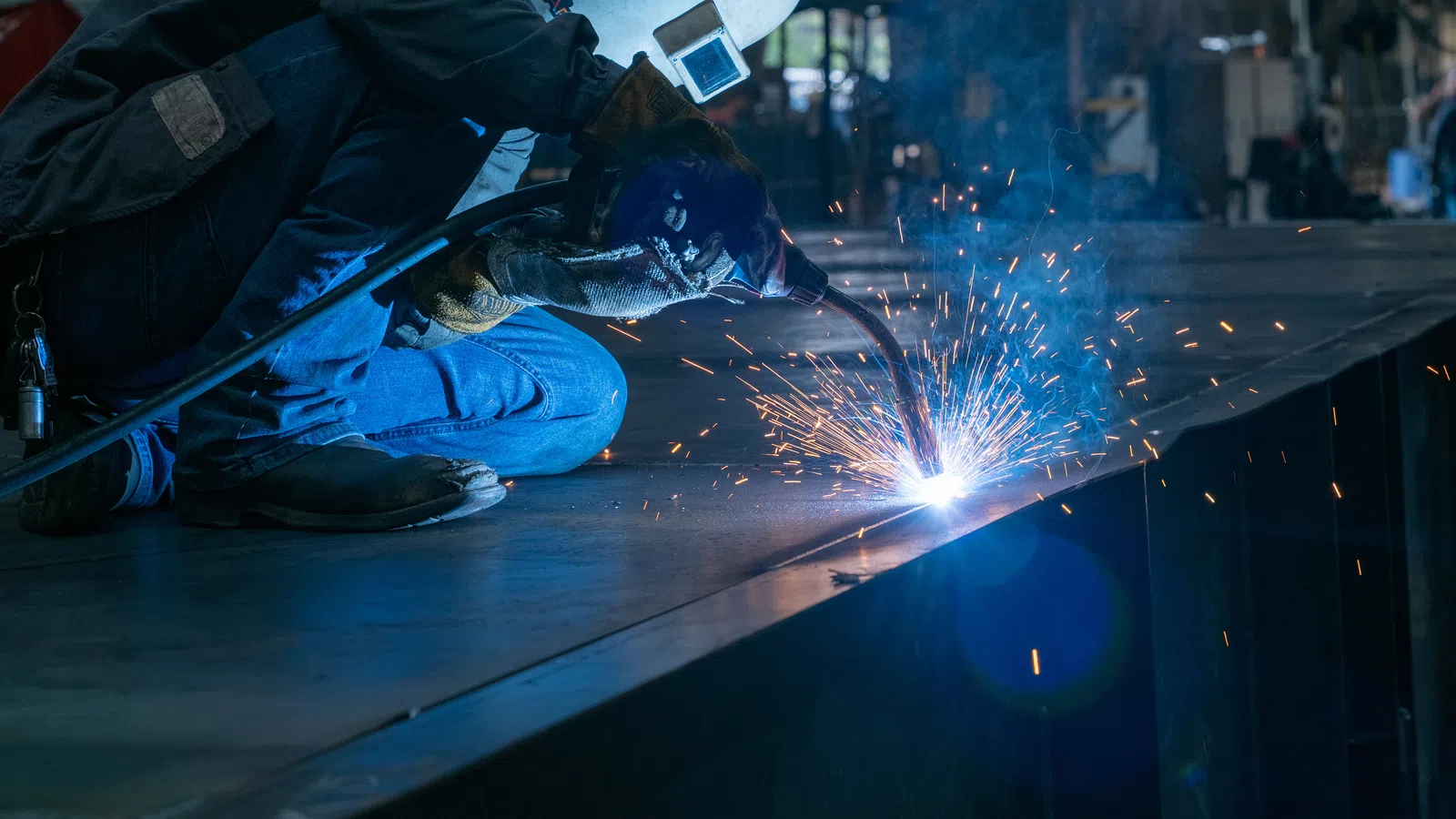 Welder in protective gear kneeling and welding metal with bright sparks in an industrial workshop.