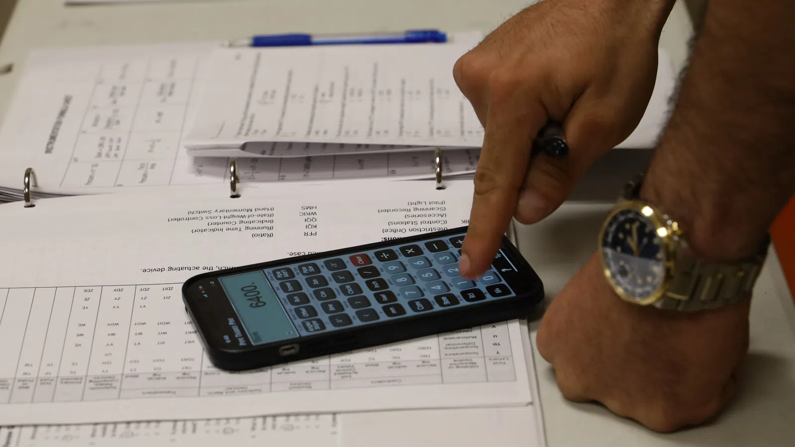 A hand using a calculator app on a smartphone with documents and a pen nearby.