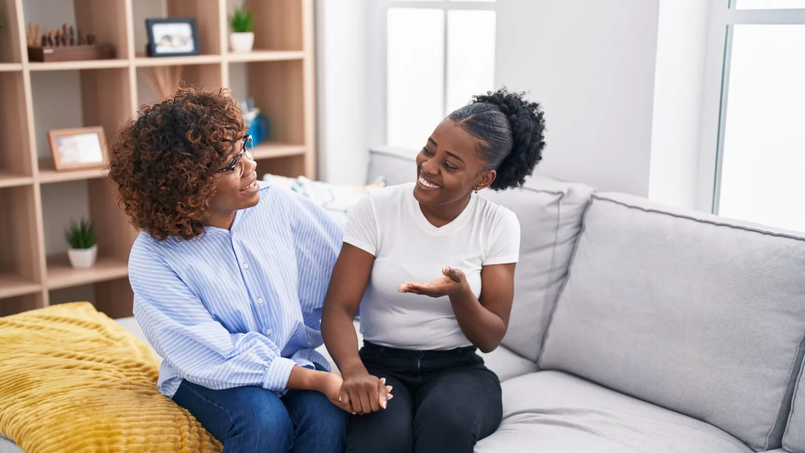 Two women sitting together on a couch, engaged in a friendly conversation and smiling.