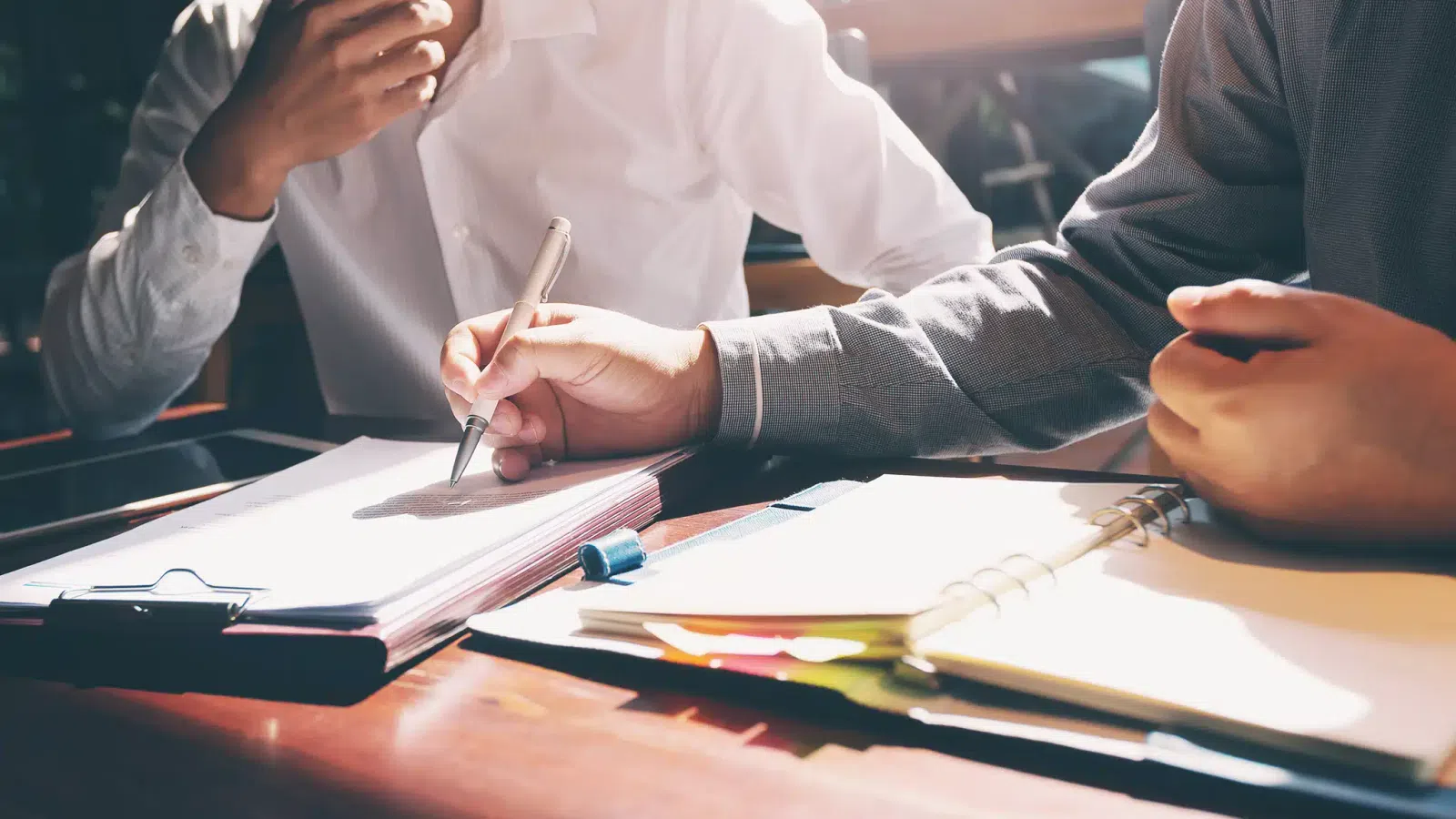 Two people working together at a desk with notebooks and pens in natural light during a meeting.