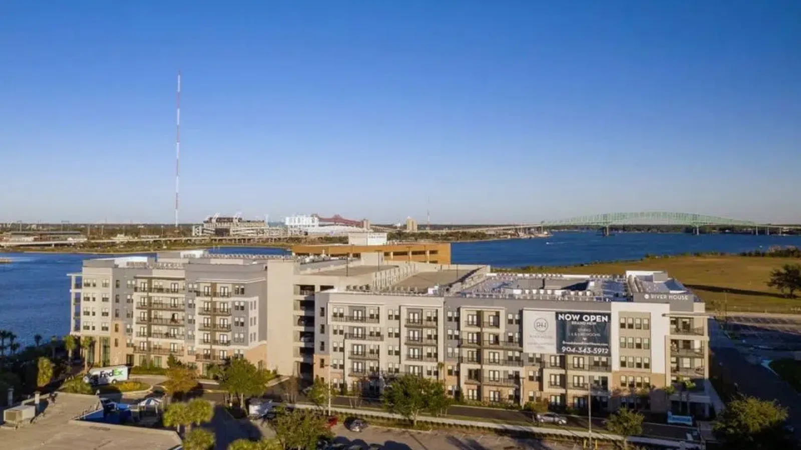Modern apartment complex by the river with a large banner, parking lot, and a green bridge in the background under a clear blue sky