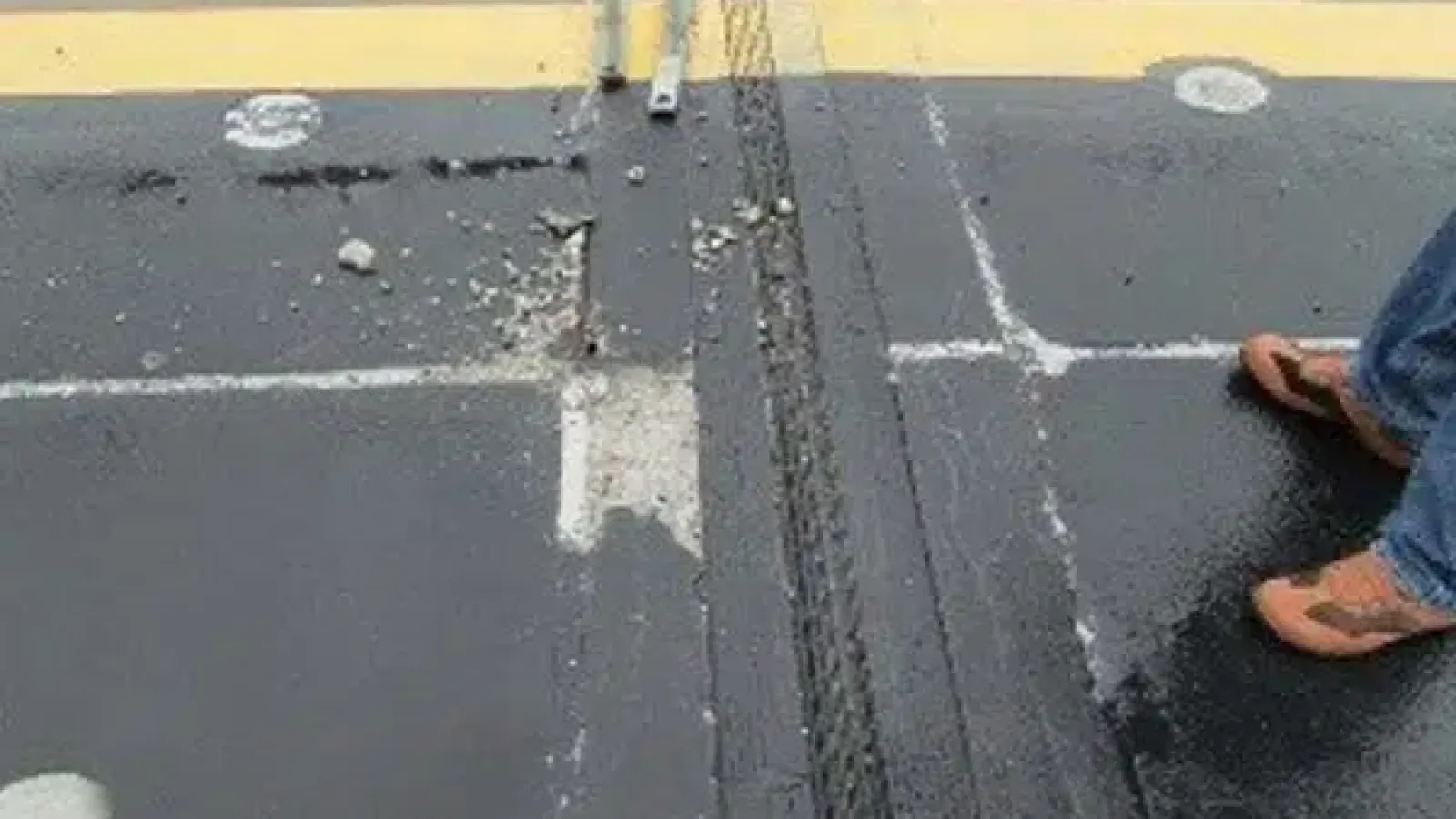 Damaged asphalt pavement with a large crack and chipped area near a pedestrian crossing with feet in sandals visible