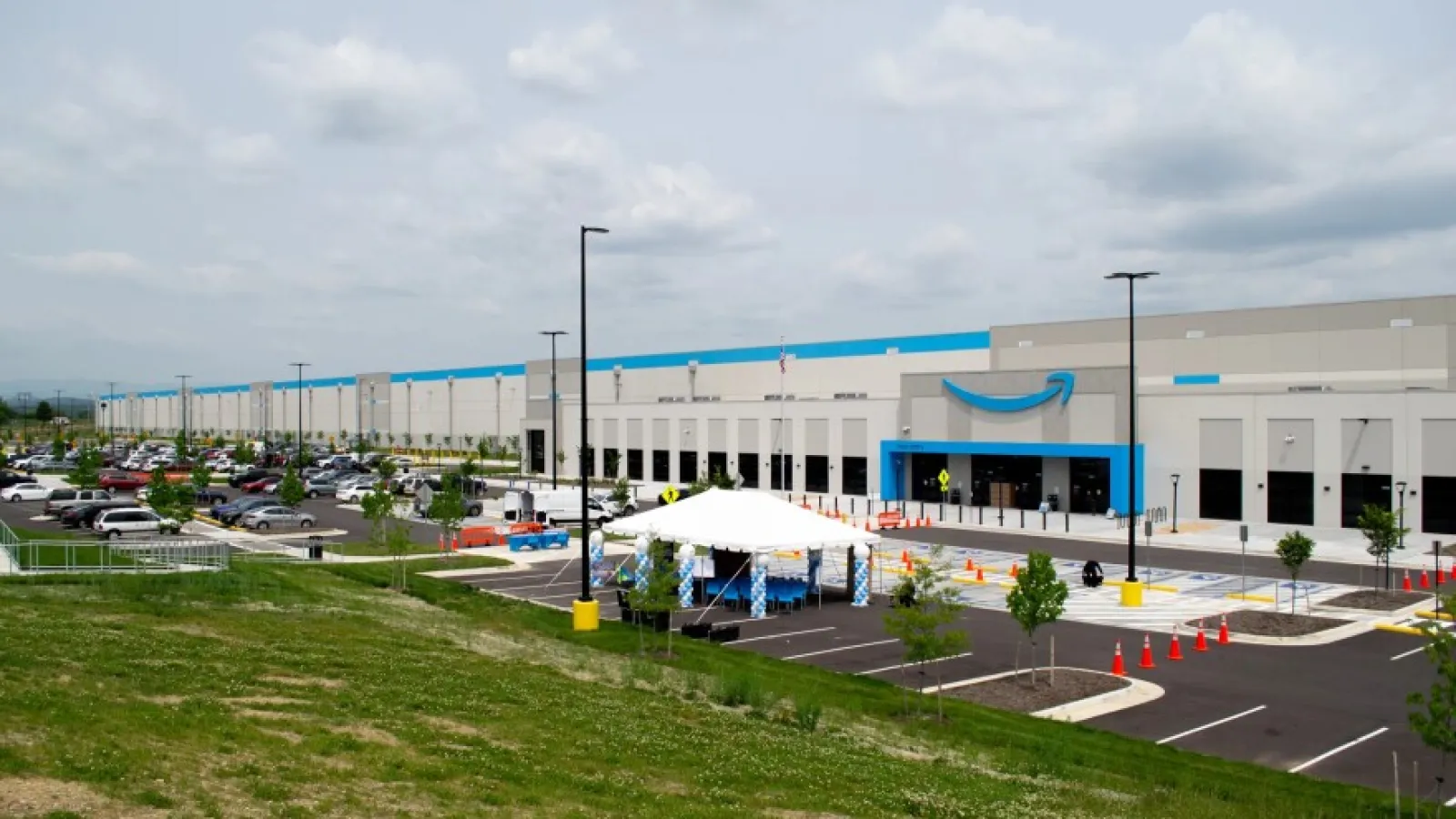 Amazon warehouse building with parking lot, cars, tents, and green grassy foreground under cloudy sky.