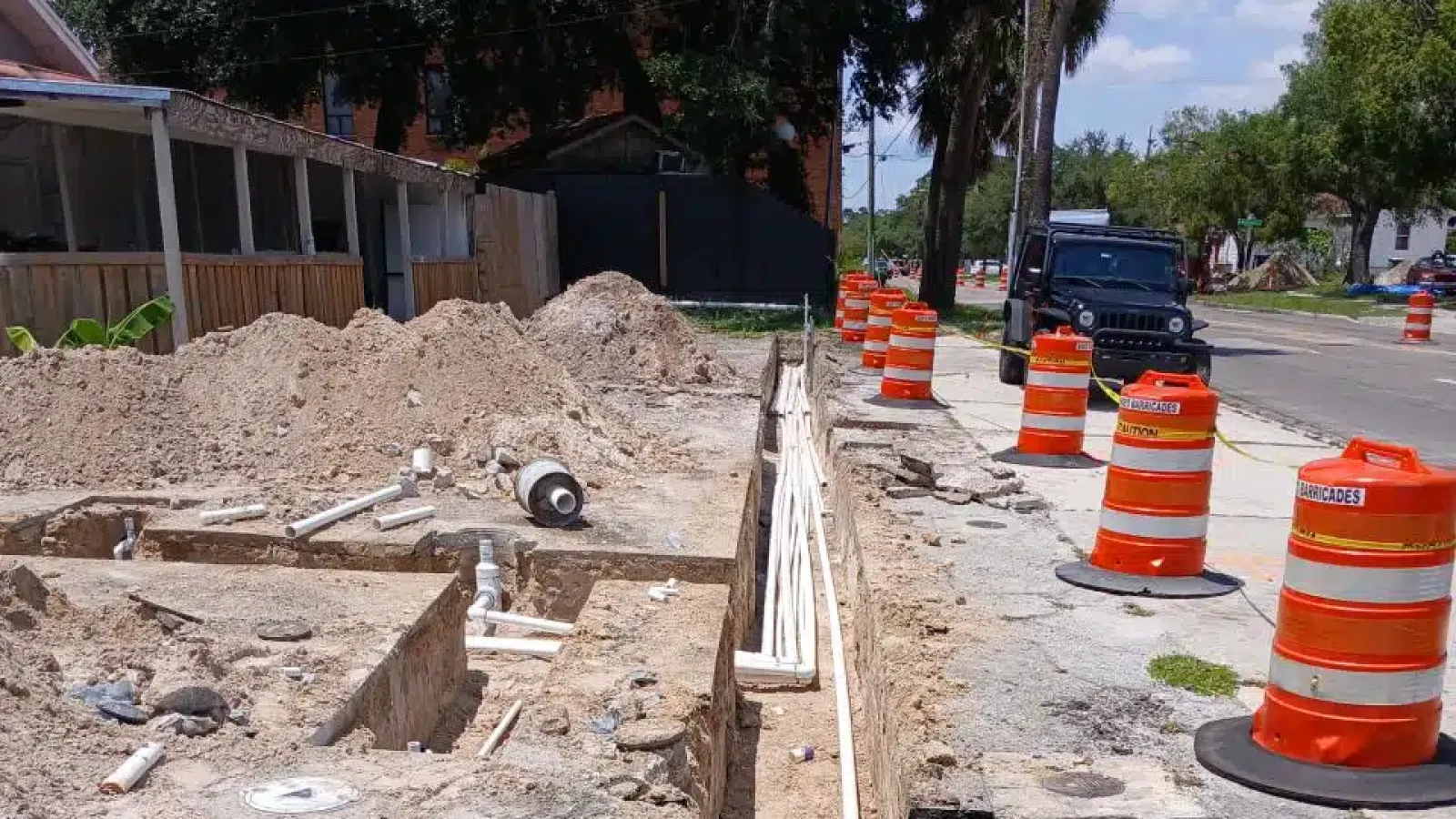 Construction site with trenches and pipes next to a street lined with orange traffic barrels on a sunny day