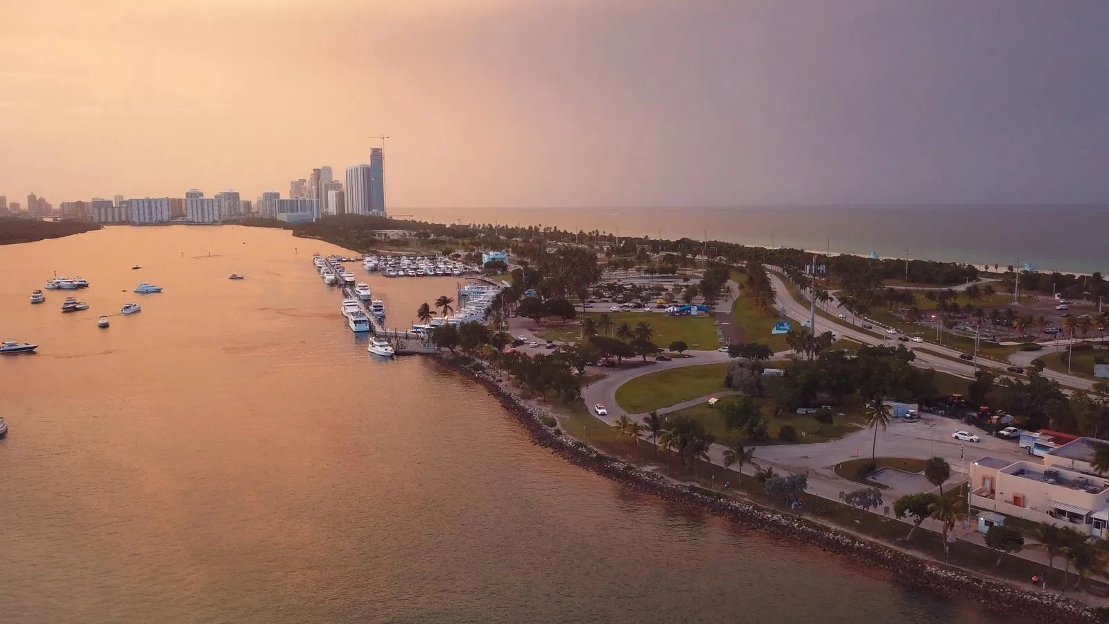 Aerial view of a coastal city at sunset with yachts docked, palm trees, high-rise buildings, and a calm ocean.