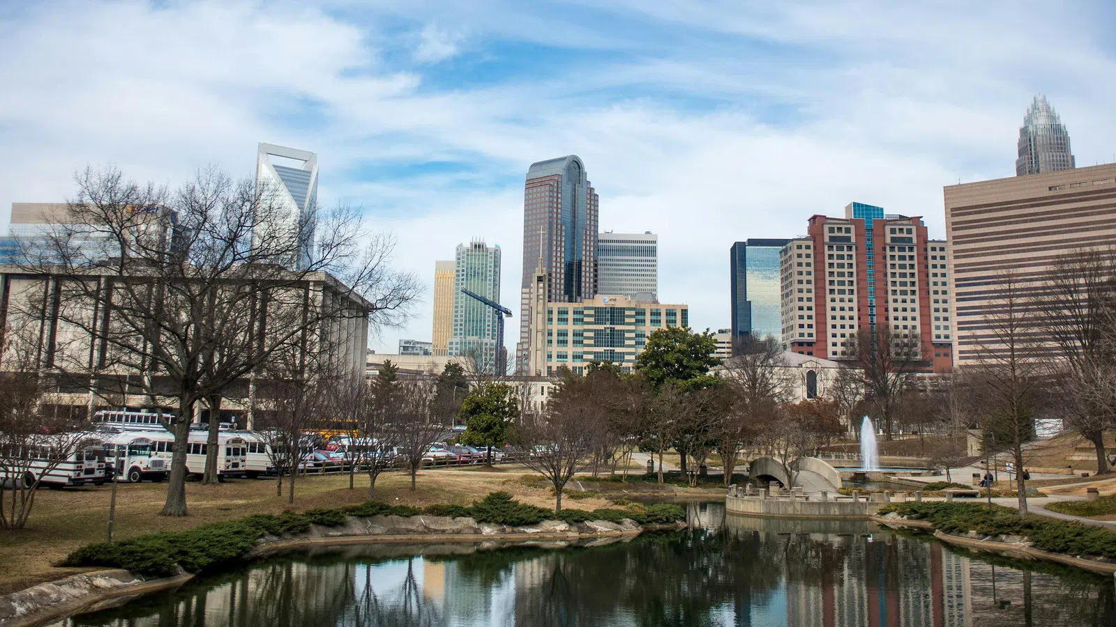 Charlotte skyline reflected in a calm urban pond with a park and cloudy blue sky above.