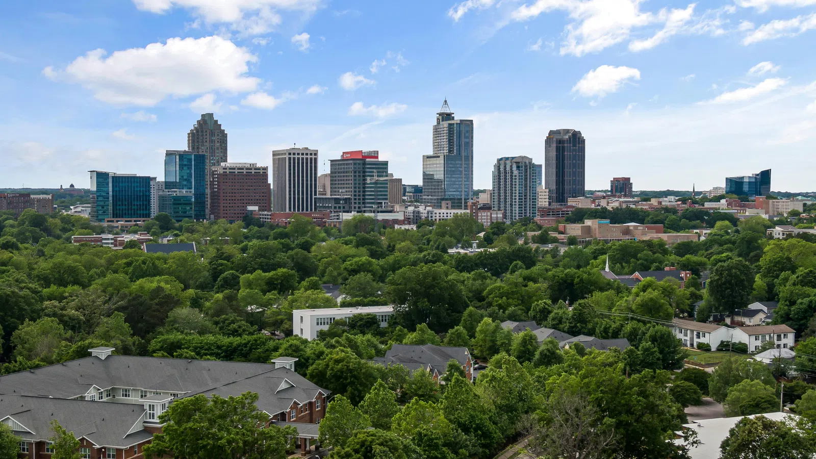 A city skyline with tall buildings surrounded by dense green trees under a bright blue sky with clouds.