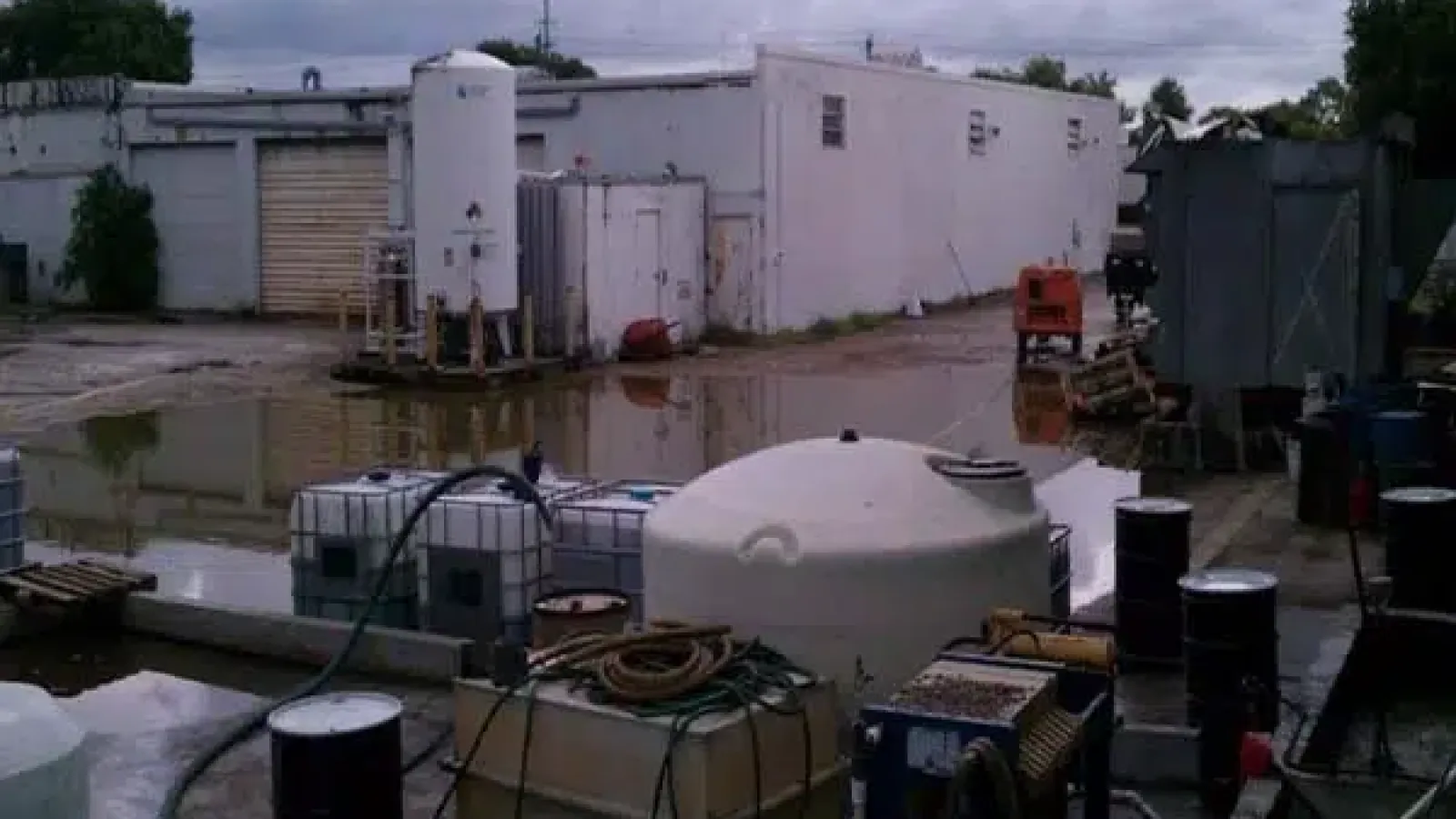 Industrial outdoor area with storage tanks, barrels, and puddles under a cloudy sky near white buildings.