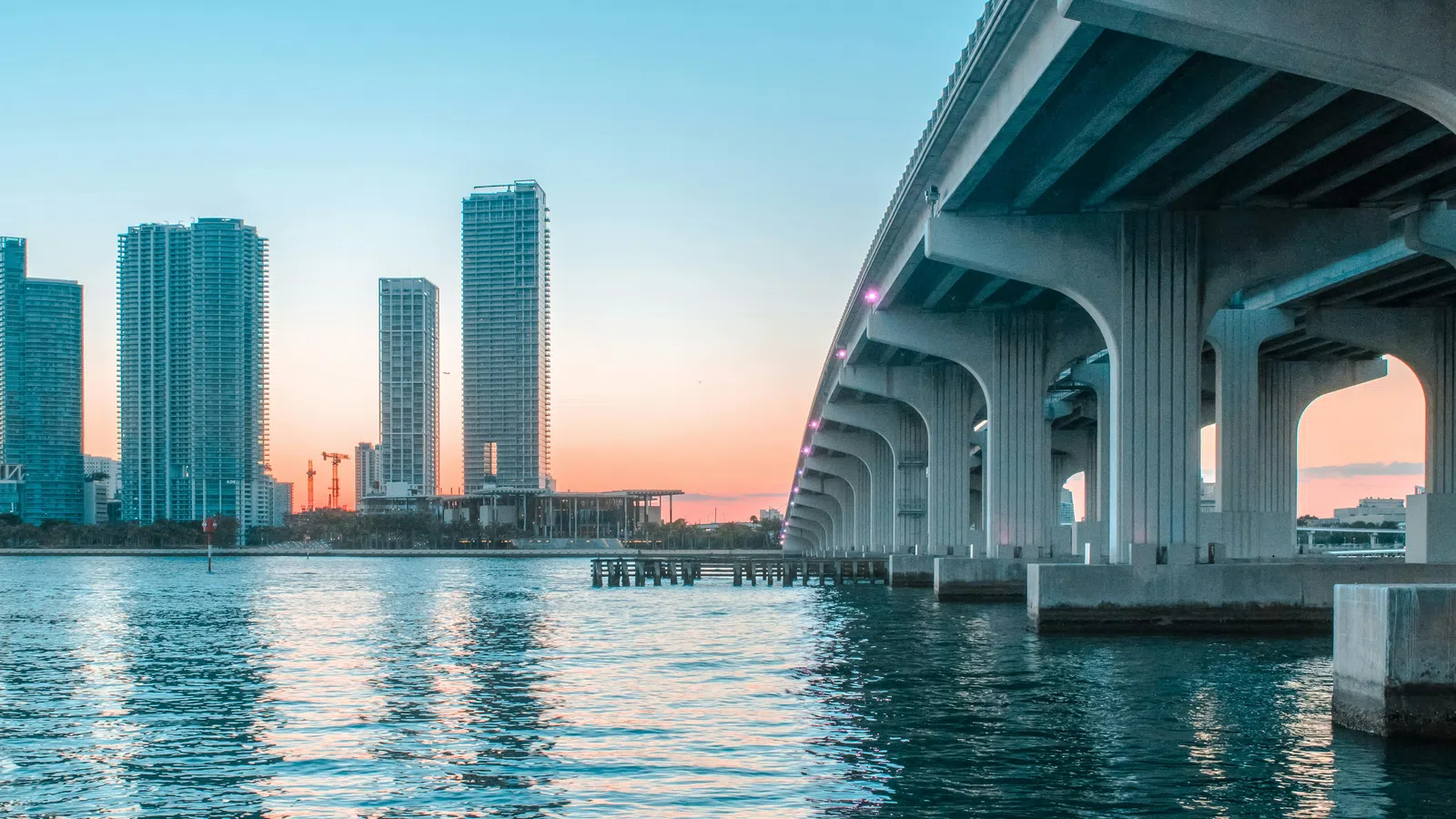 Sunset city skyline with high-rise buildings and bridge over calm waterfront reflecting colorful sky.