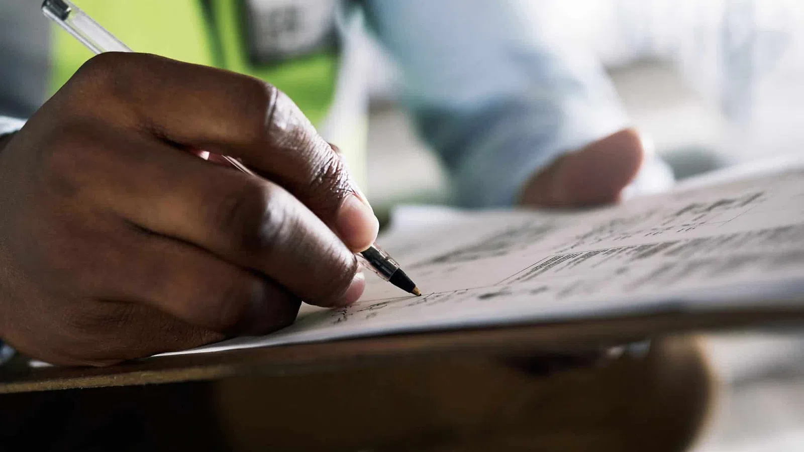 Worker in safety vest writing notes on clipboard with pen at construction site or warehouse