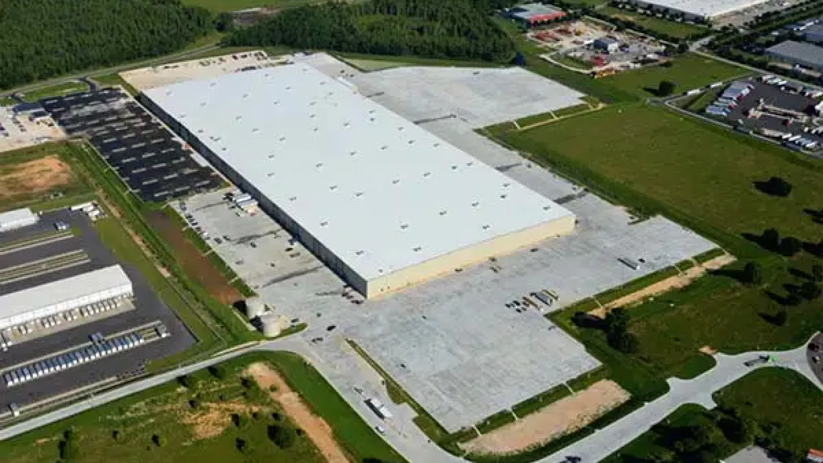 Aerial view of a large industrial warehouse surrounded by parking lots and green fields under a clear sky.