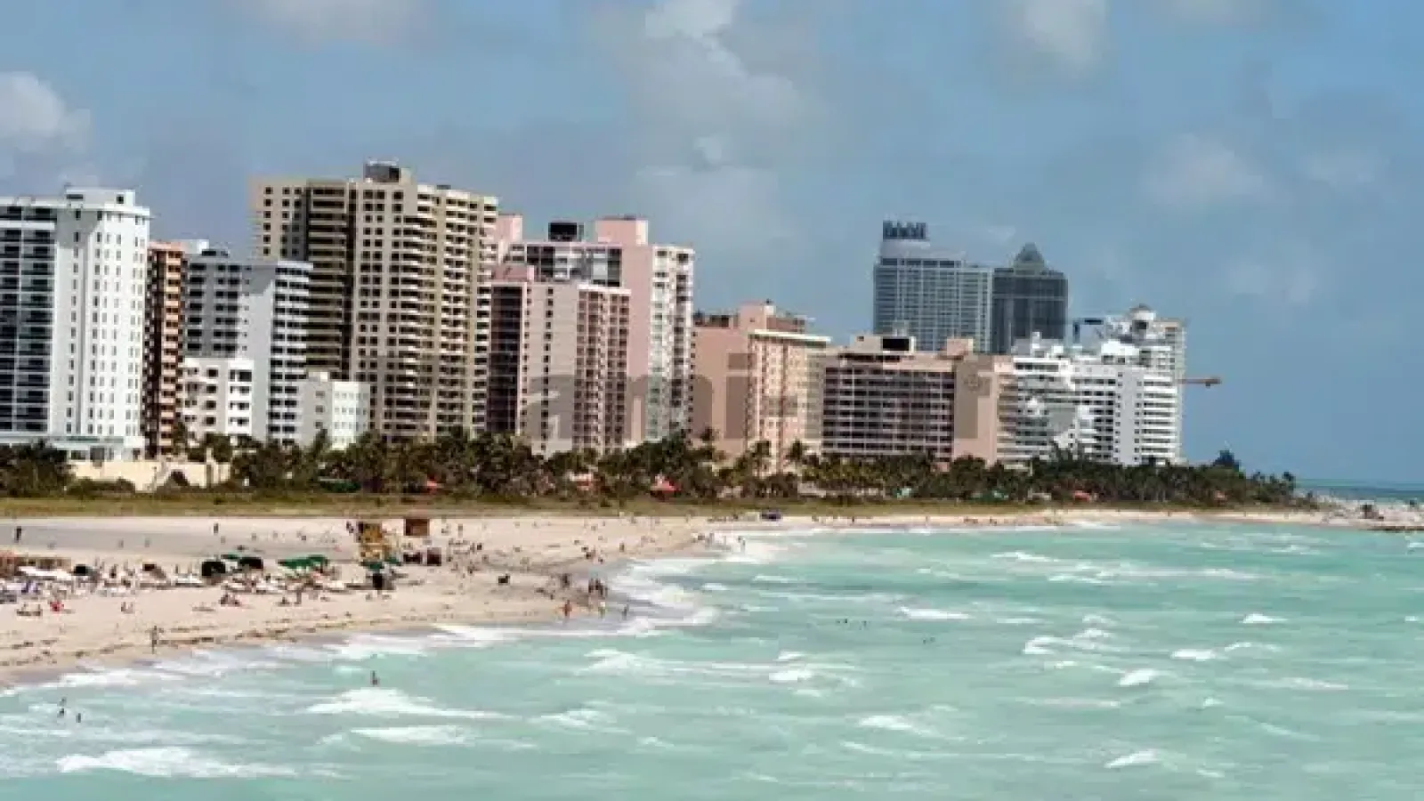 Sunny beach with turquoise waves, sandy shore, palm trees, and high-rise buildings along the coastline