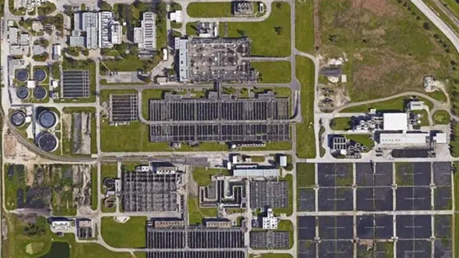 Aerial view of a large wastewater treatment plant with multiple rectangular ponds, tanks, and buildings on green land.