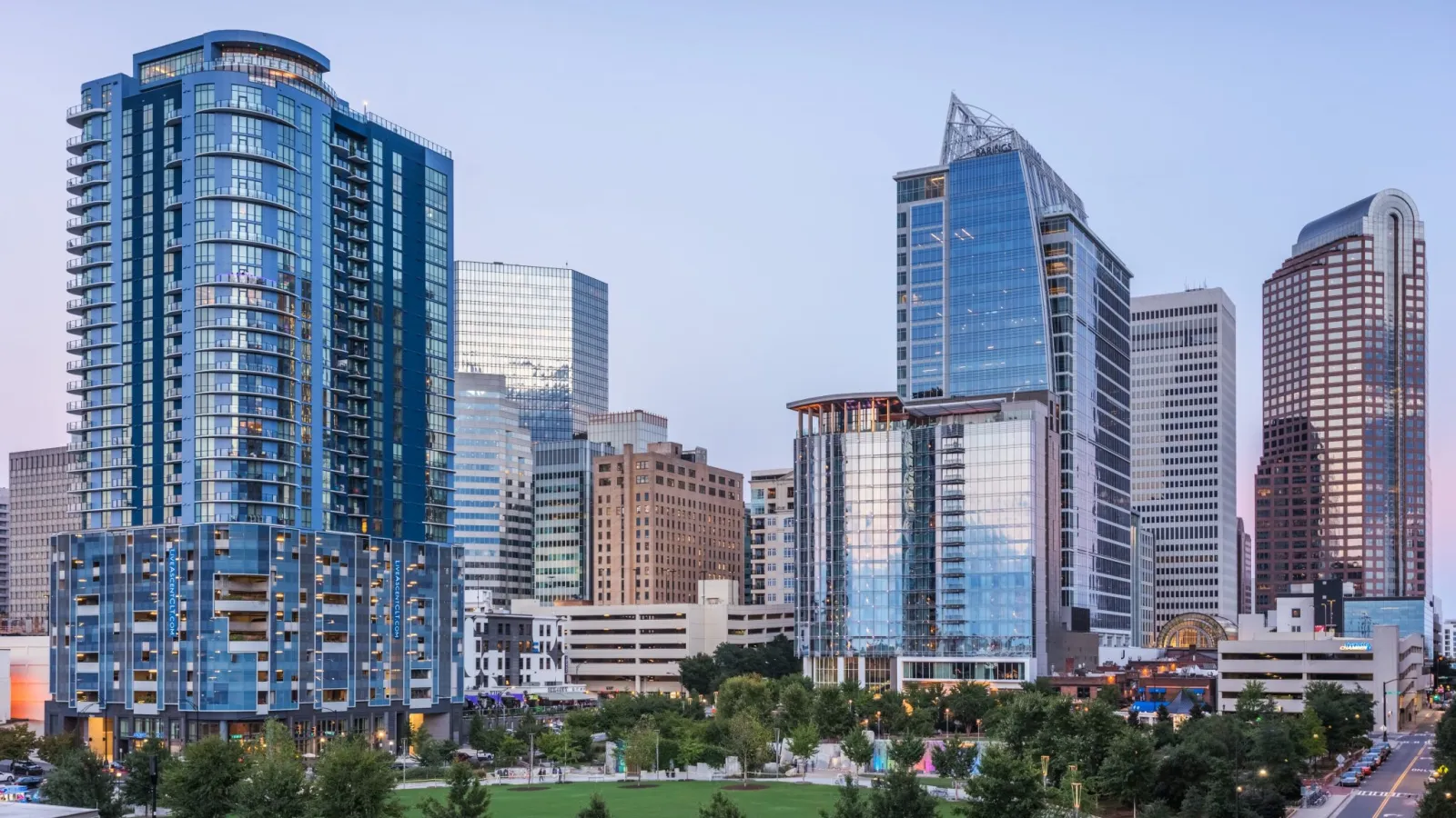 City skyline with modern skyscrapers and green park under a clear blue sky at dusk.