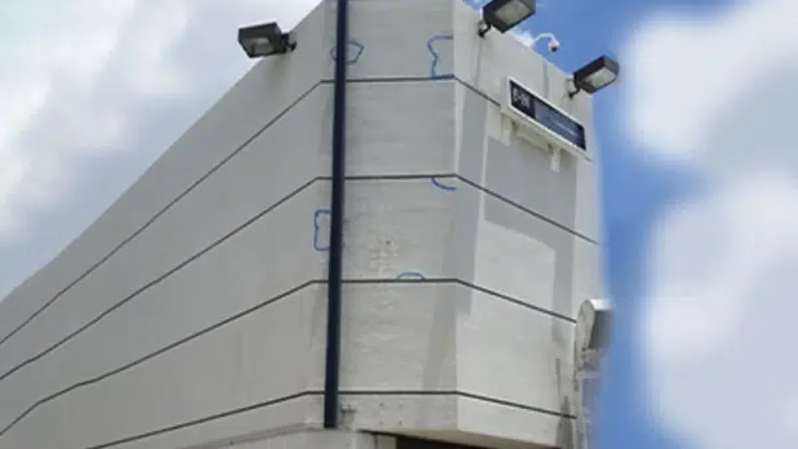 White concrete building corner with external lights under a partly cloudy blue sky.