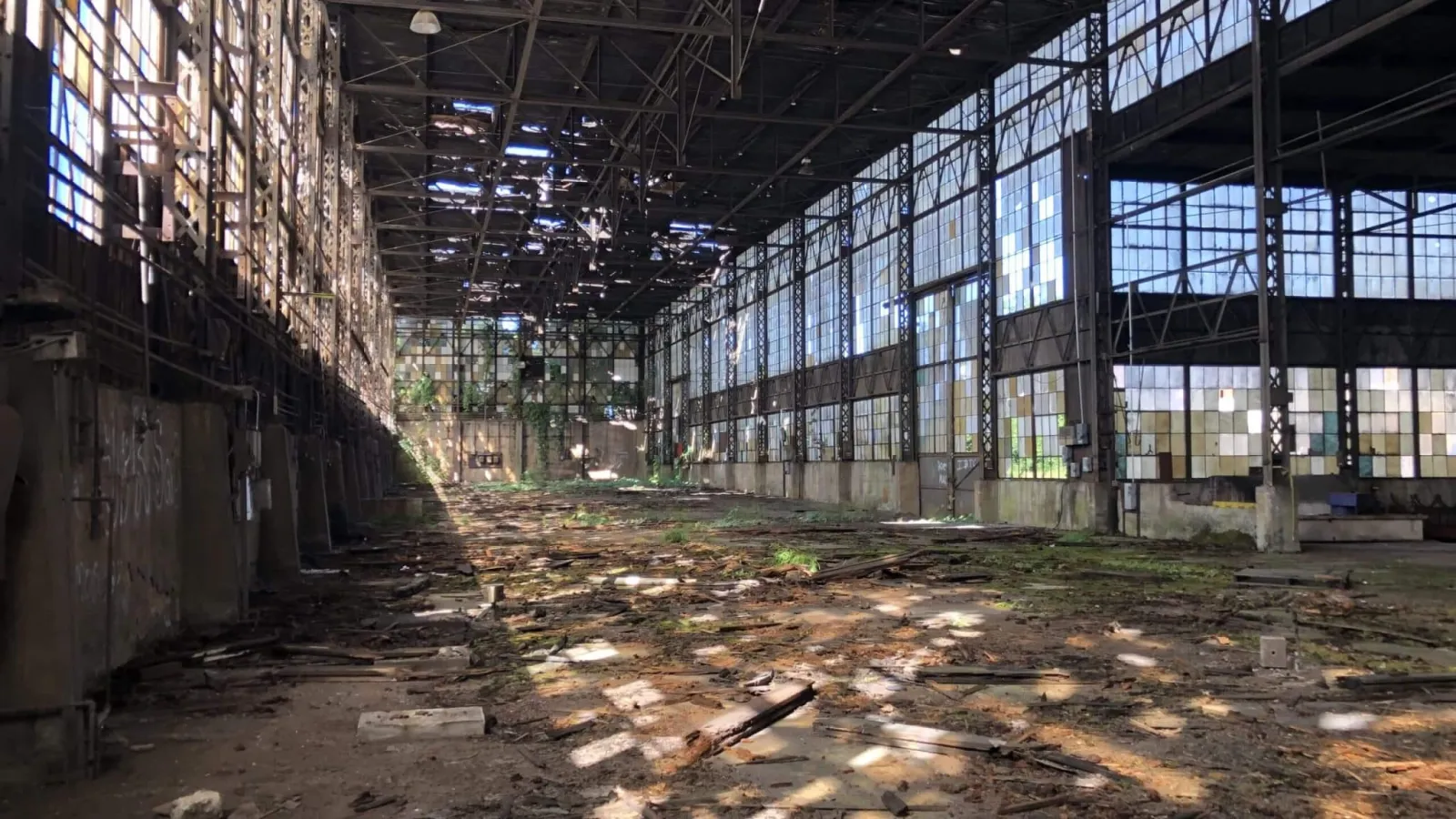 Interior of a large abandoned warehouse with broken windows and debris scattered on the floor.