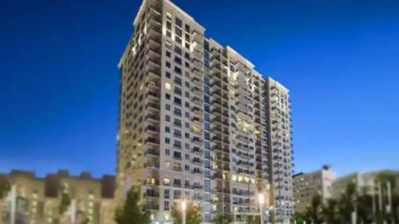 Tall residential apartment building lit up at dusk with clear blue sky and surrounding cityscape.