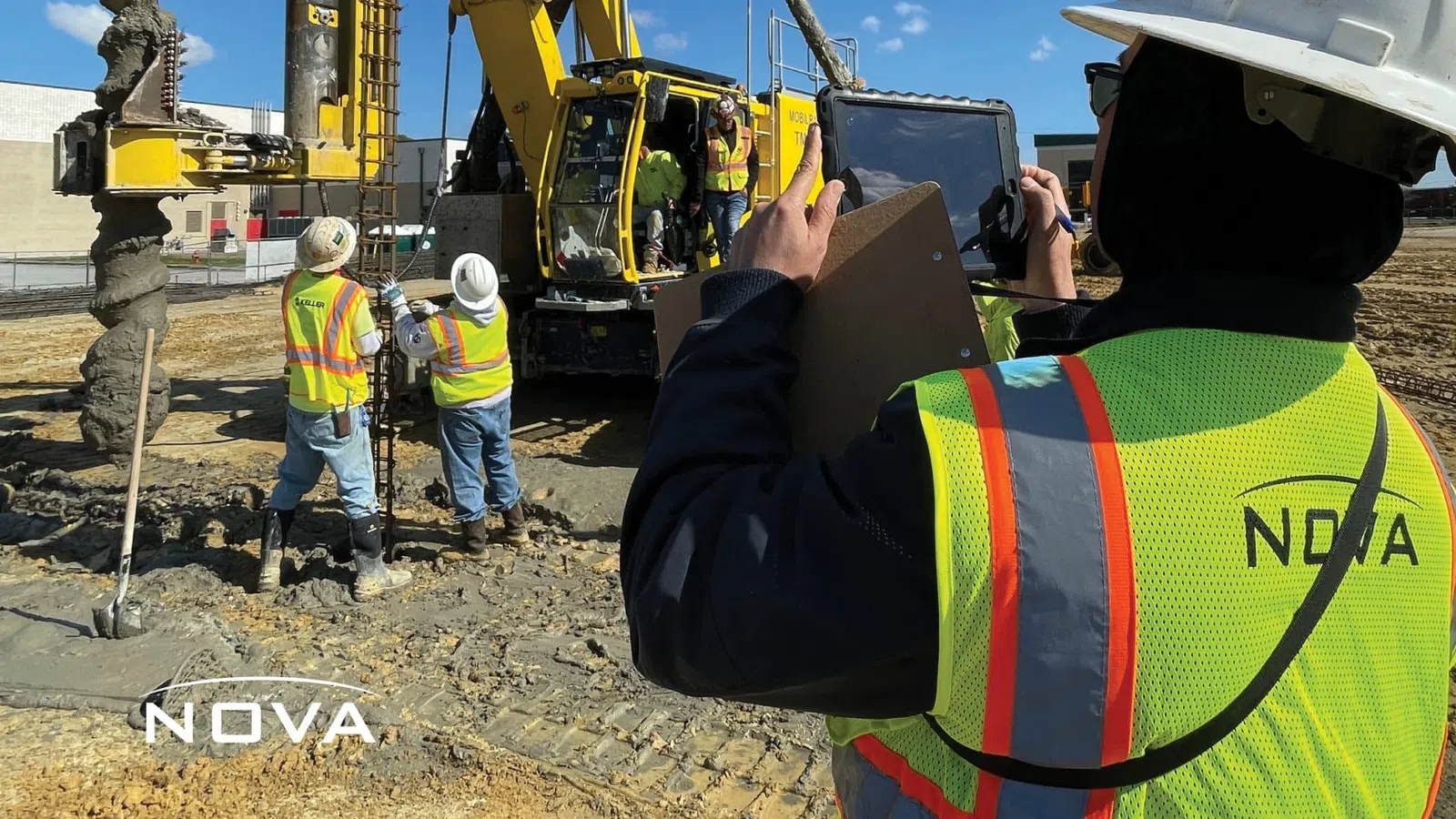 Construction workers operate heavy drilling machinery on-site while a supervisor documents progress using a tablet.