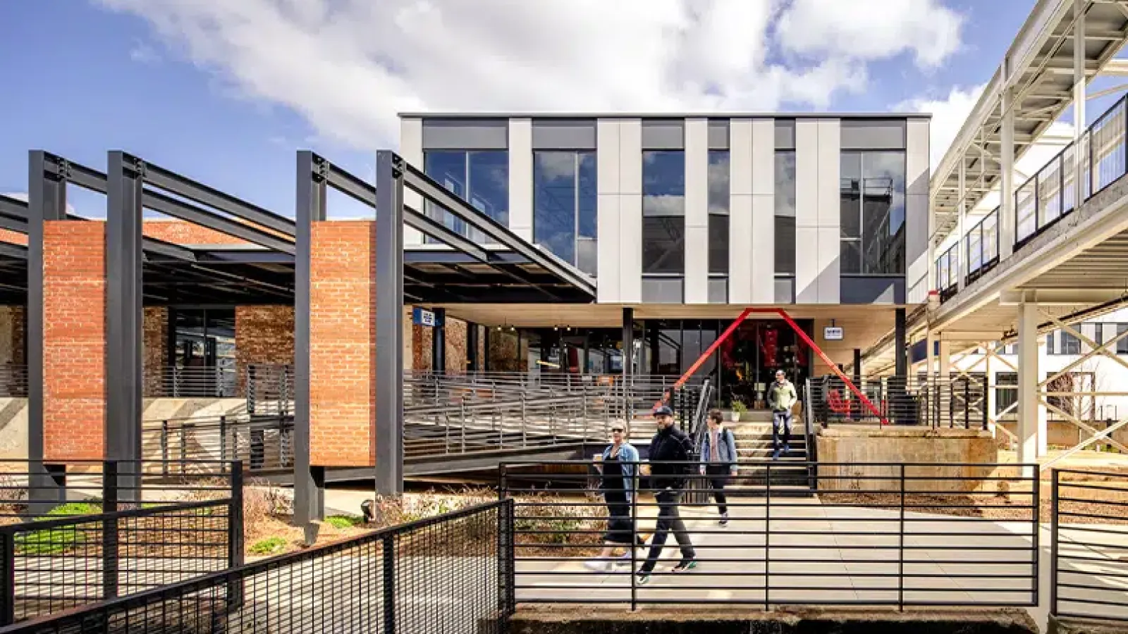 Modern architectural building with glass, brick, and metal elements and people walking on pathway under blue sky.