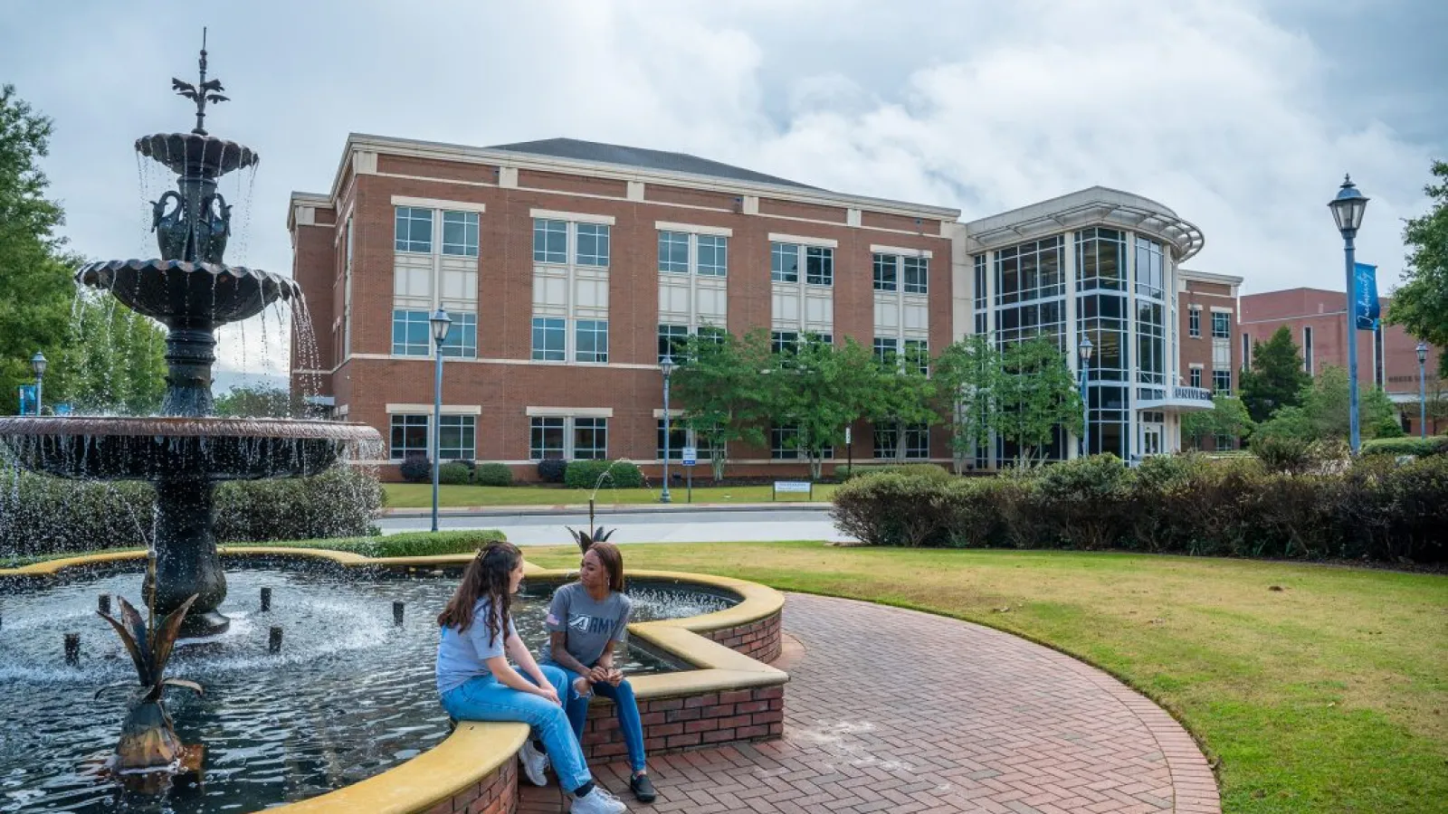 Two students sit by a large water fountain on a brick path outside a modern university building on a cloudy day