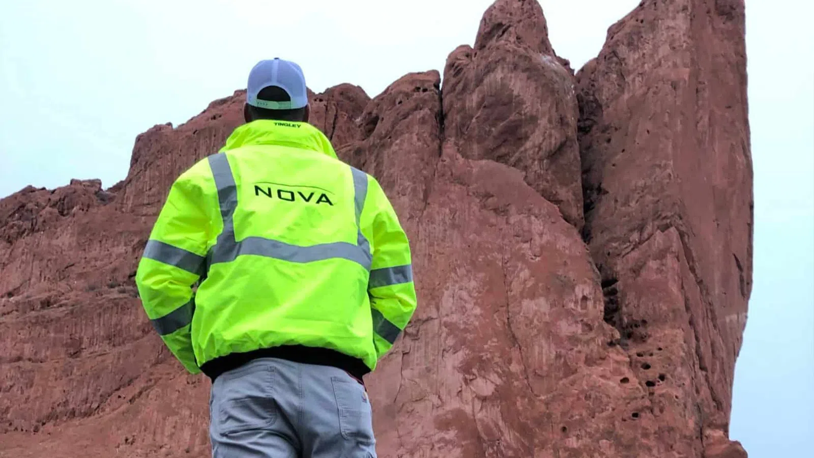 Person wearing a bright yellow safety jacket labeled NOVA stands facing large red rock formations under a clear sky.