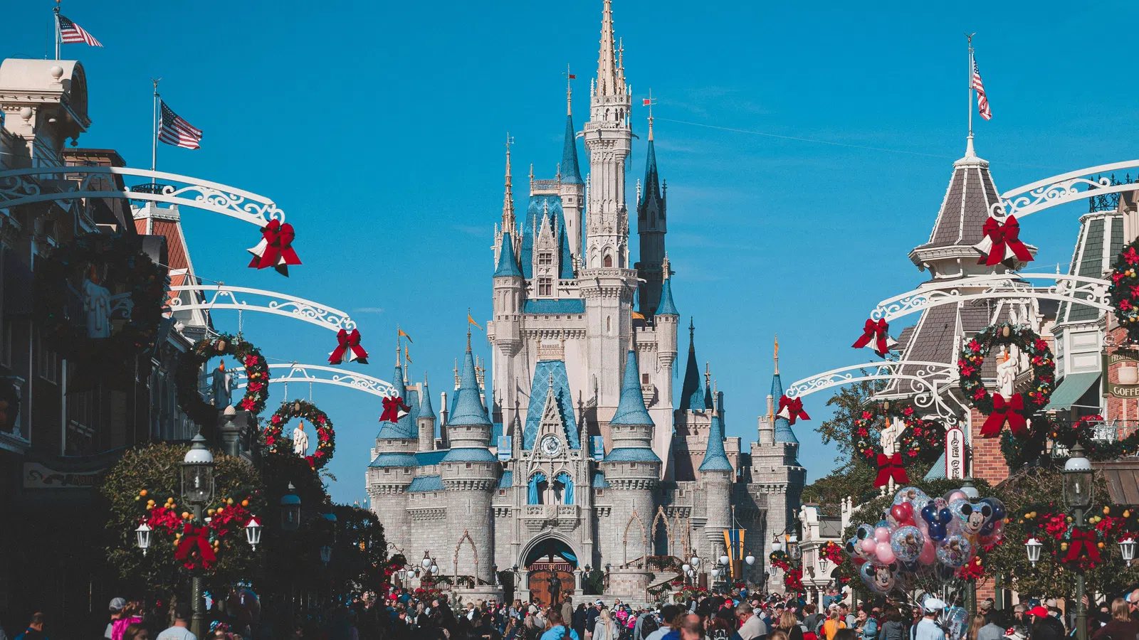 Disney castle in bright daylight with holiday wreaths and bows lining Main Street packed with visitors.