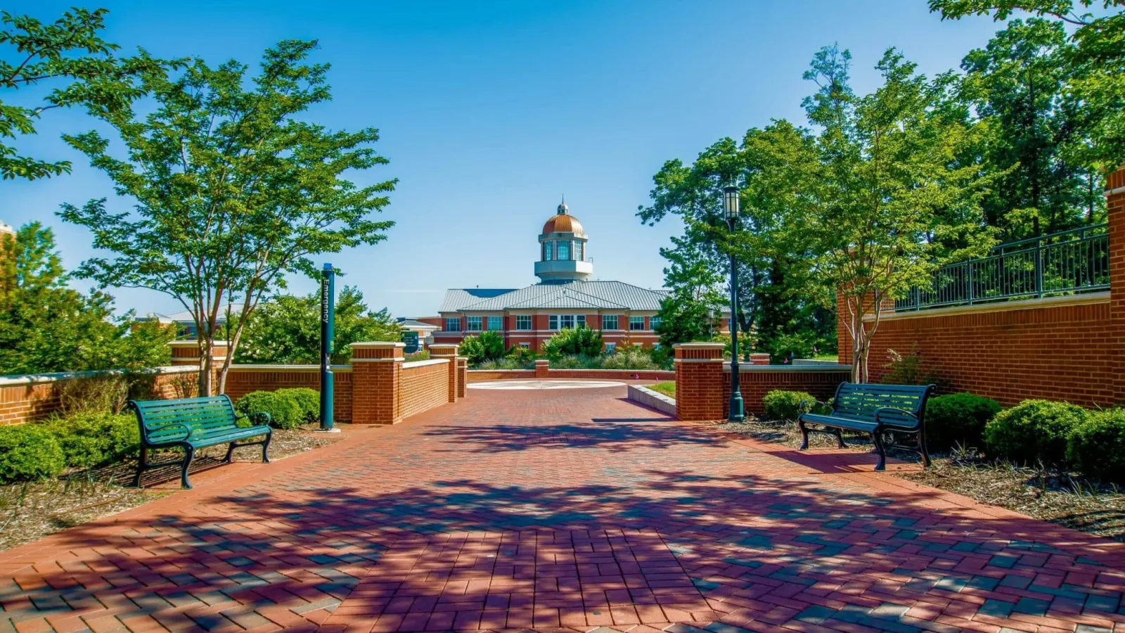 Brick pathway with benches and trees leading to a building with a domed cupola under a clear blue sky.